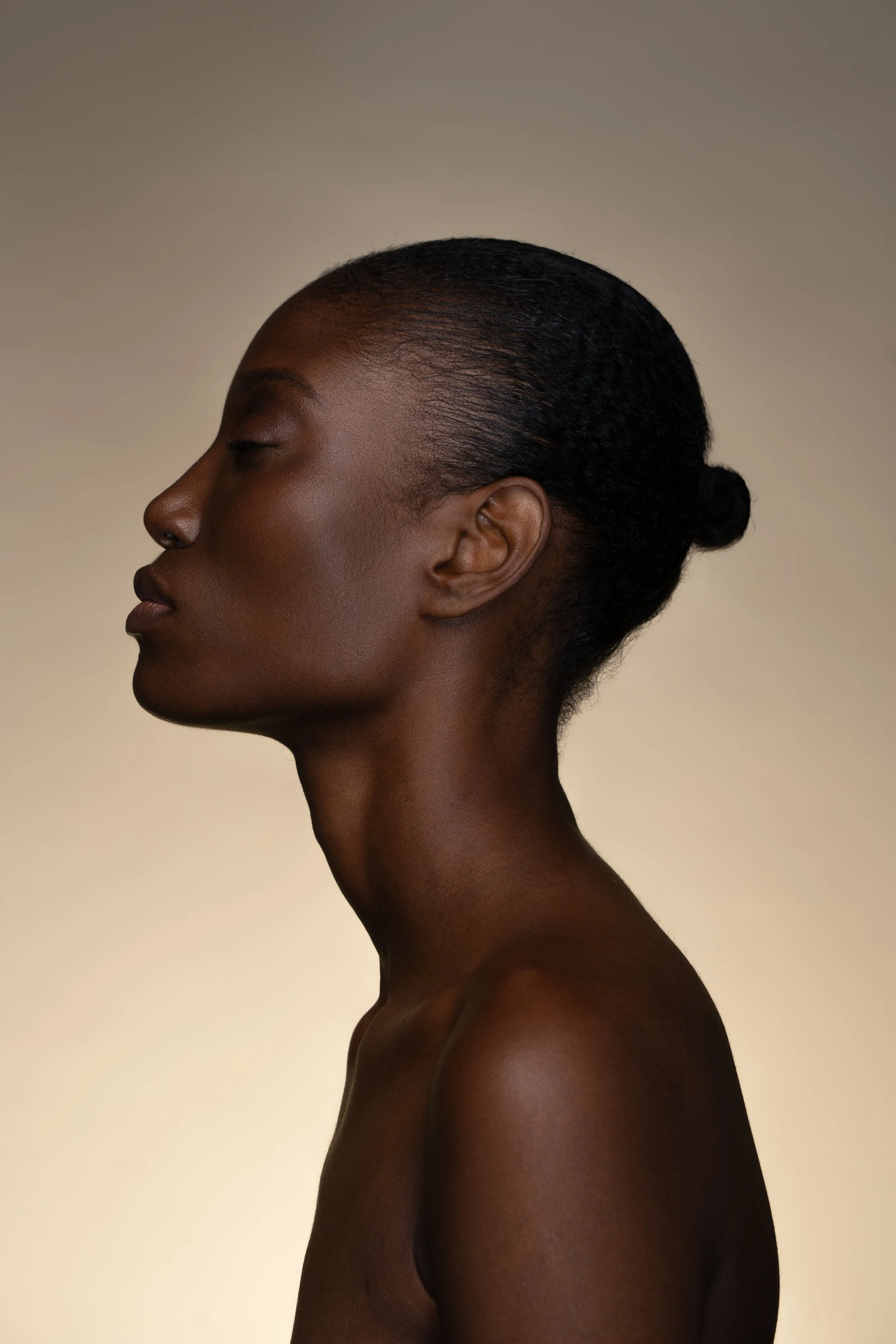 Profile view of a woman with dark skin and closed eyes, her hair pulled back into a bun, against a beige gradient background.
