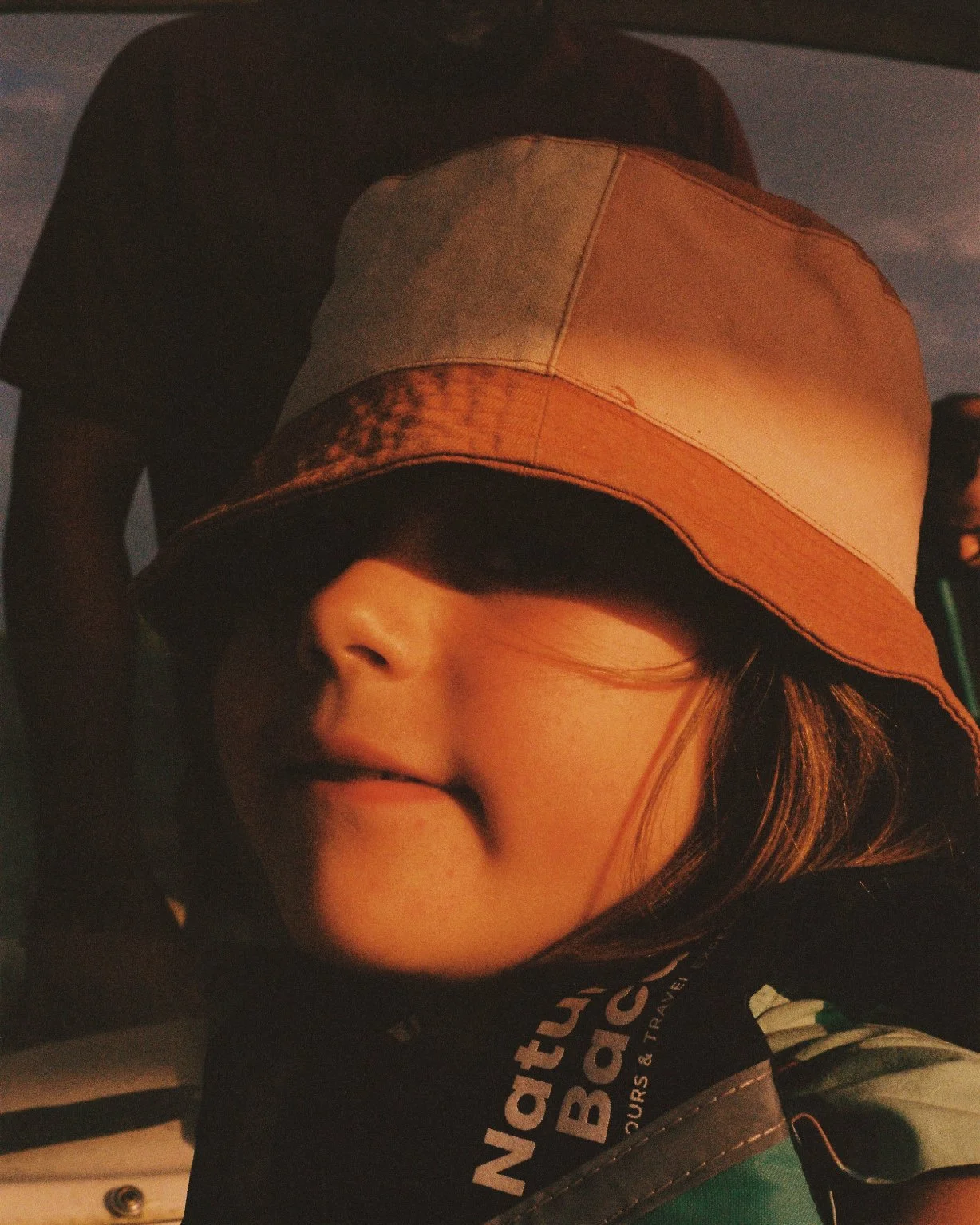 A young girl wearing a tan and brown bucket hat, smiling with her mouth slightly open, and a black and green jacket, with some text visible on the jacket.