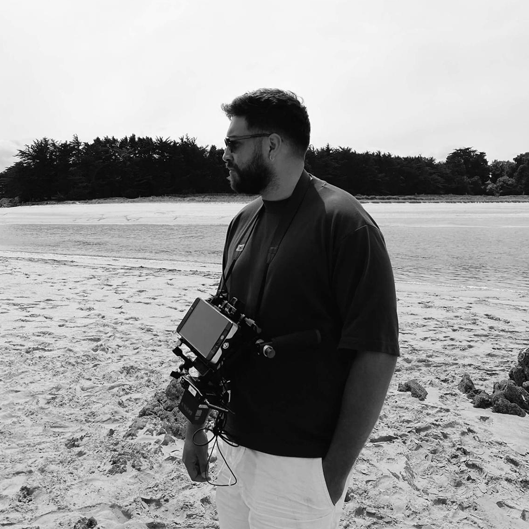 A man with a beard and sunglasses standing on a sandy beach, holding a camera with a screen, wearing a dark t-shirt and light-colored pants, with trees in the background.