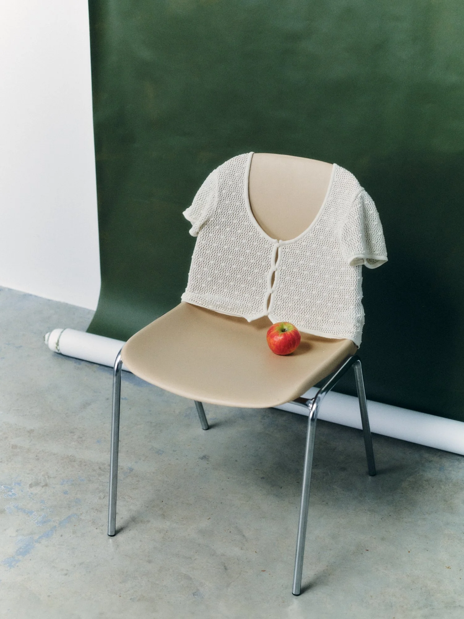 The image features an empty beige chair with a cream-colored knitted short-sleeve top draped over it. There is a red apple on the seat of the chair. The background consists of a dark green backdrop with a white roll or paper on the floor near the cha