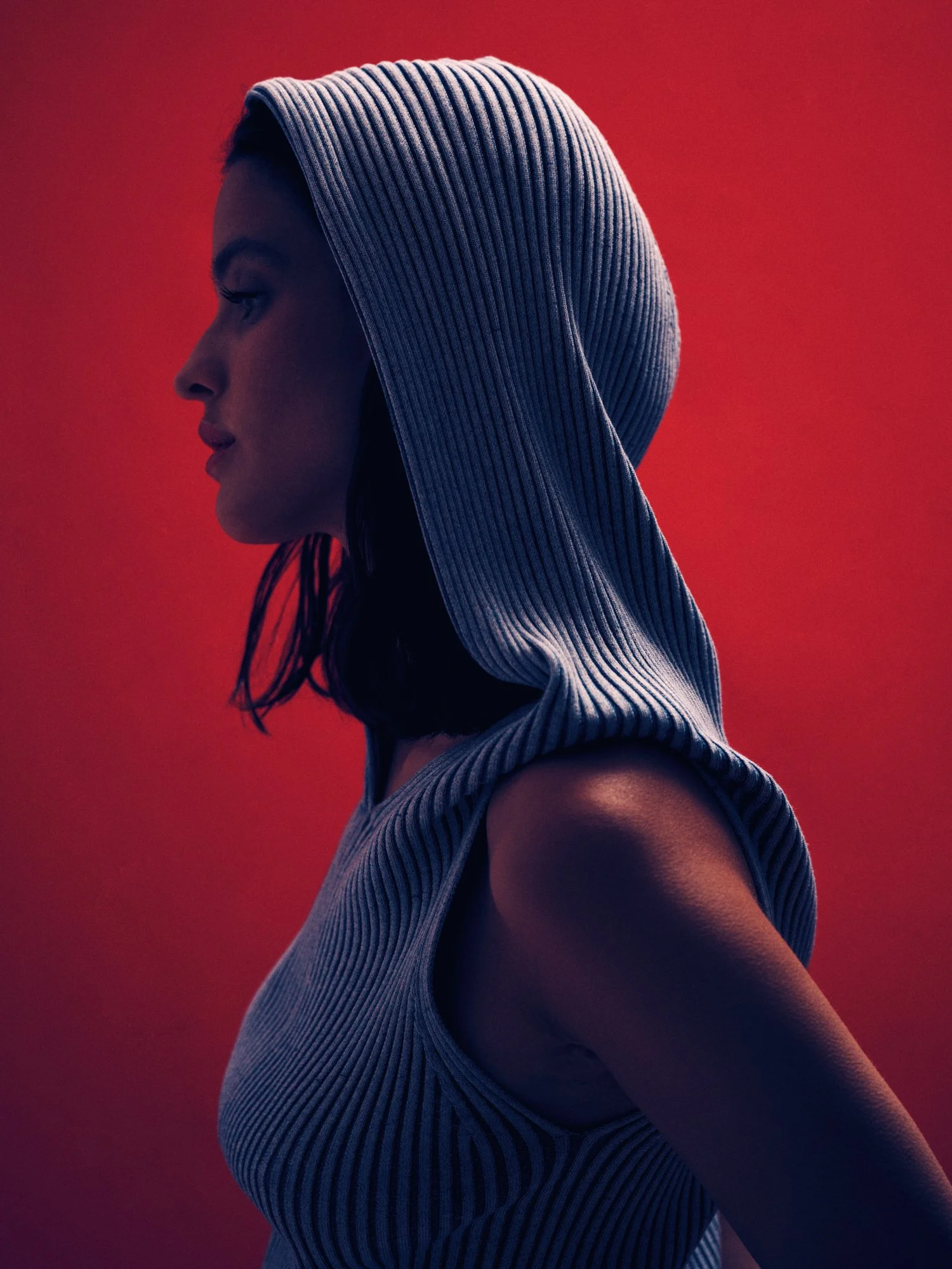 Side profile of a woman with short dark hair wearing a blue and gray striped hooded sleeveless top against a red background.