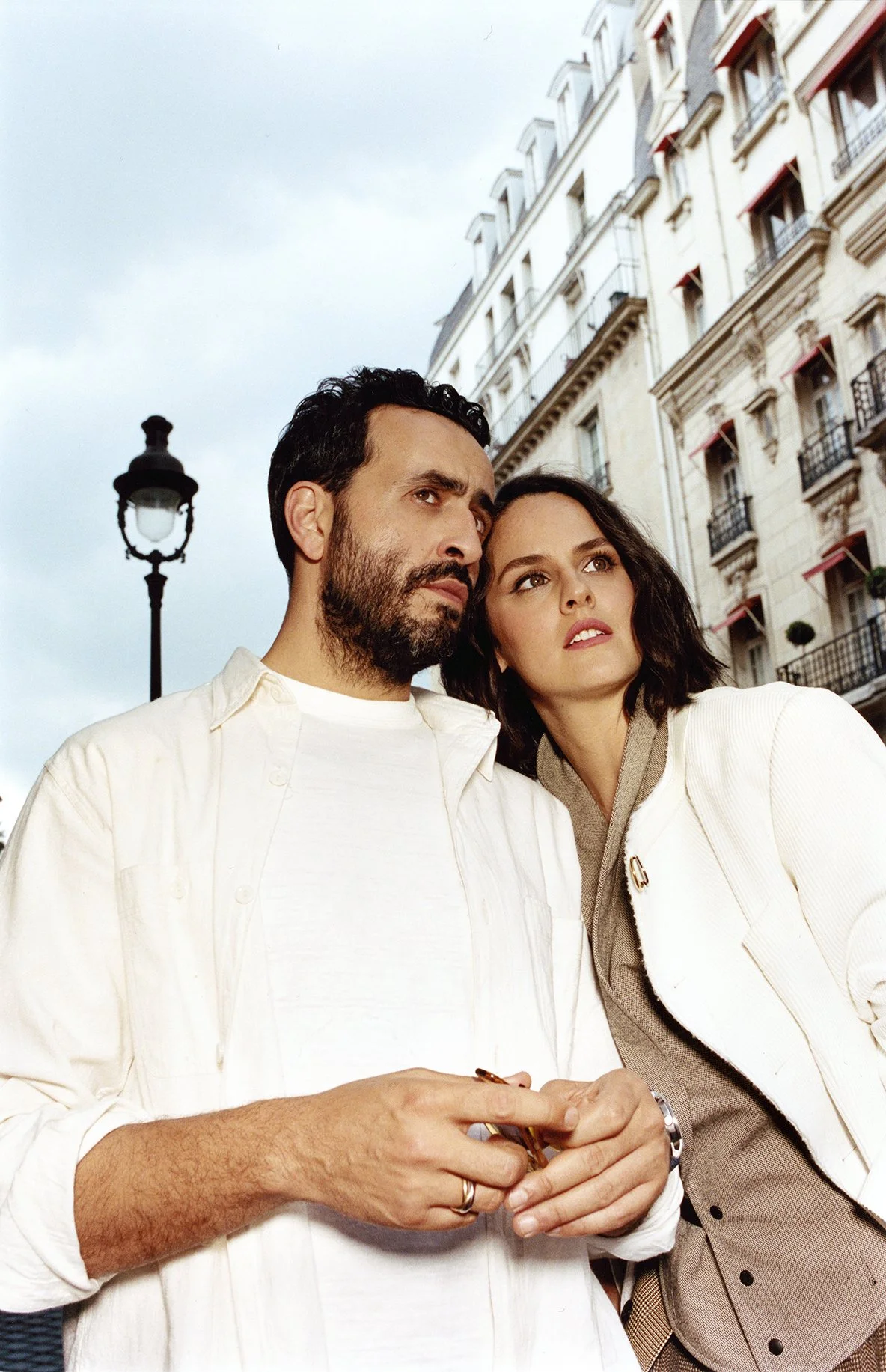 A man and a woman standing close together outdoors in front of European-style buildings, looking to the side with serious expressions.