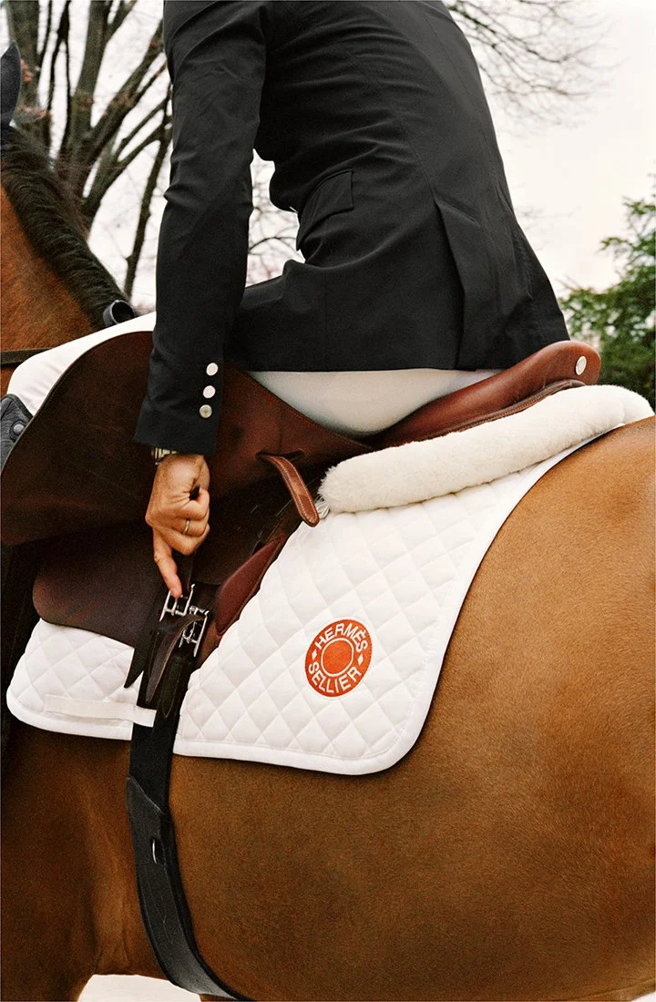 Close-up of a person wearing a black jacket on a horse with a white saddle pad labeled "Hermes Seller" and a brown leather saddle, outdoors with trees in the background.