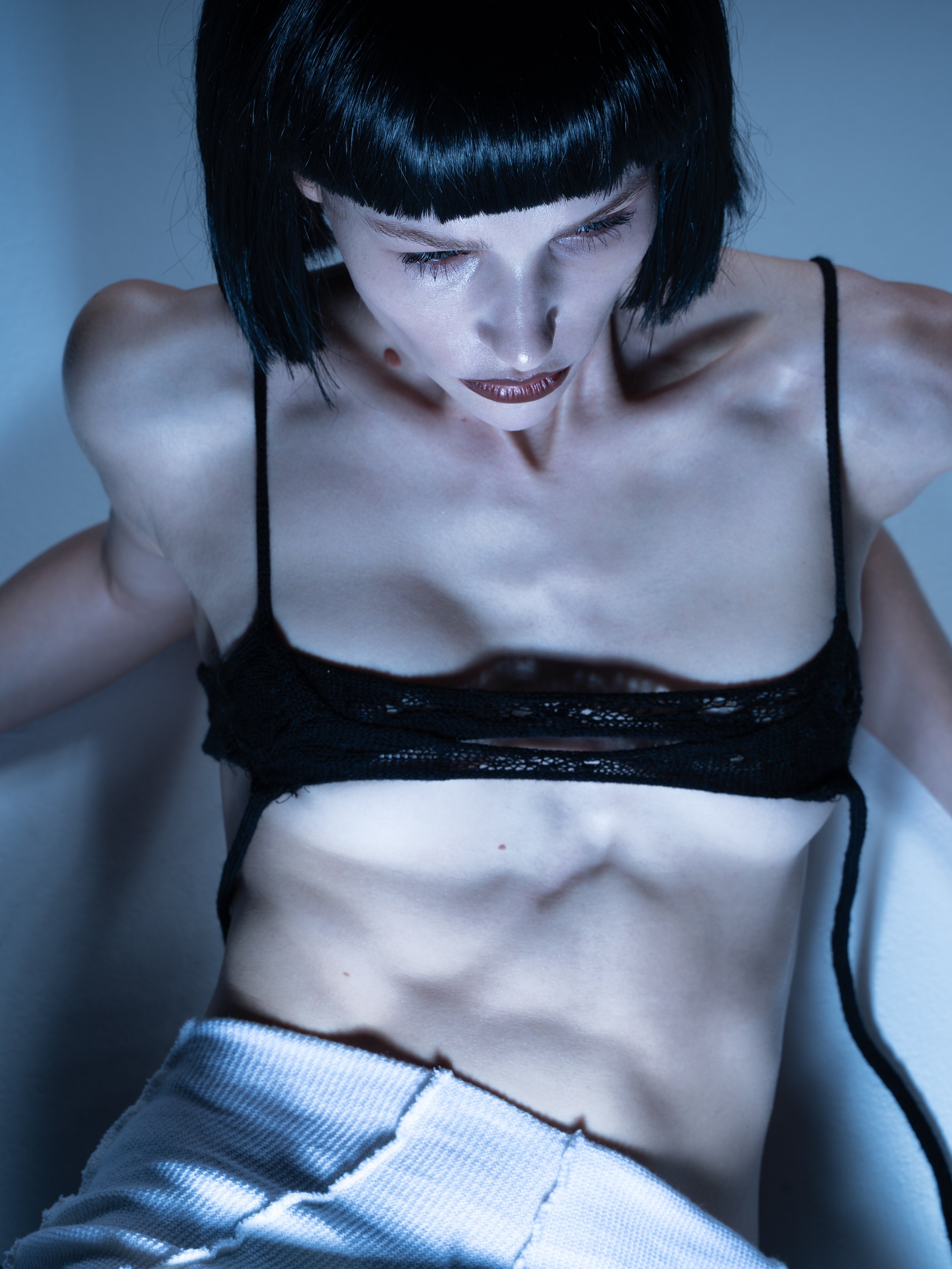 A woman with black hair and bangs looking downward, wearing a black lace top and grey pants.