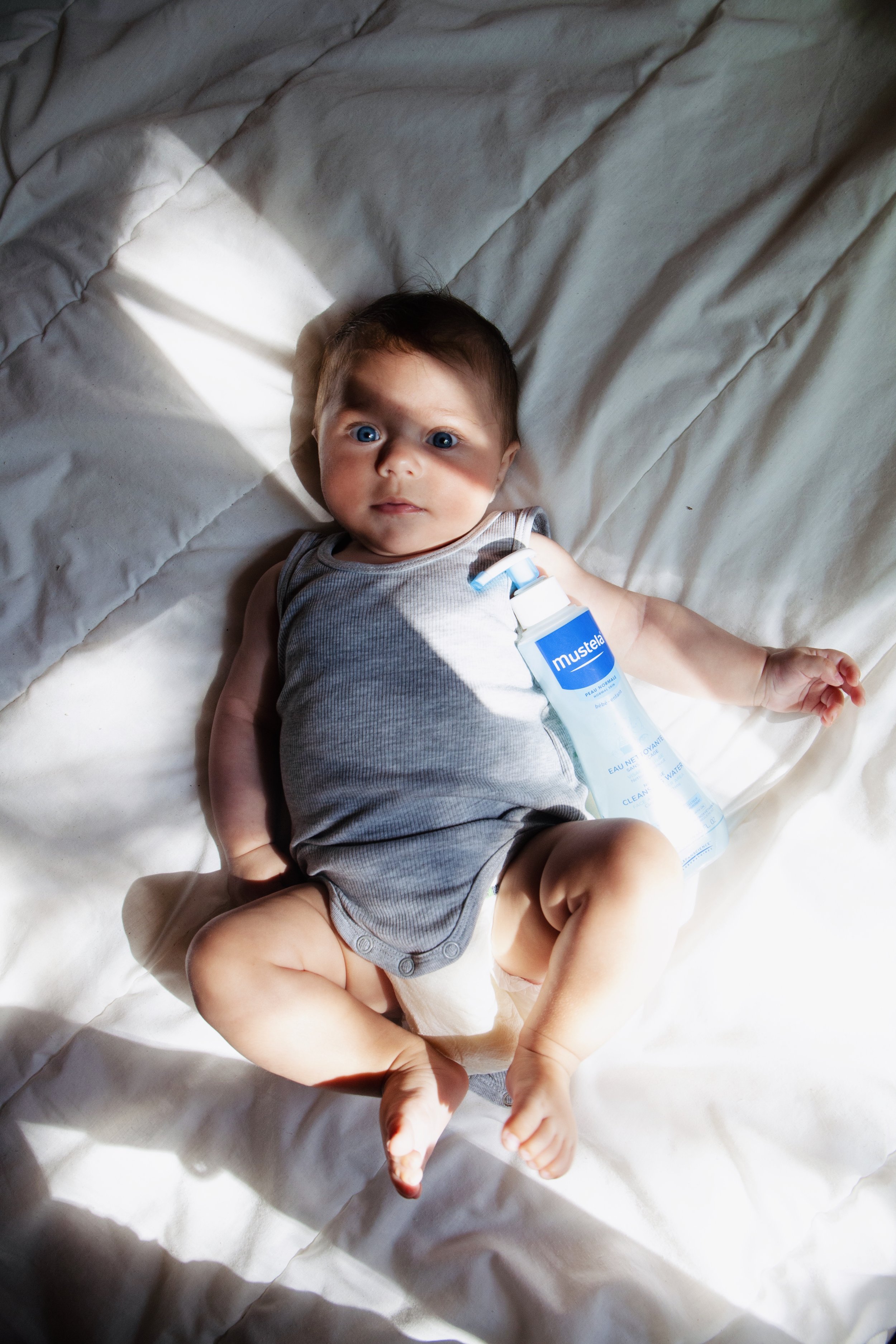 A baby lying on a white sheet, looking up, with sunlight casting shadows. The baby is wearing a gray sleeveless onesie and has a bottle of Mustela baby water beside them.