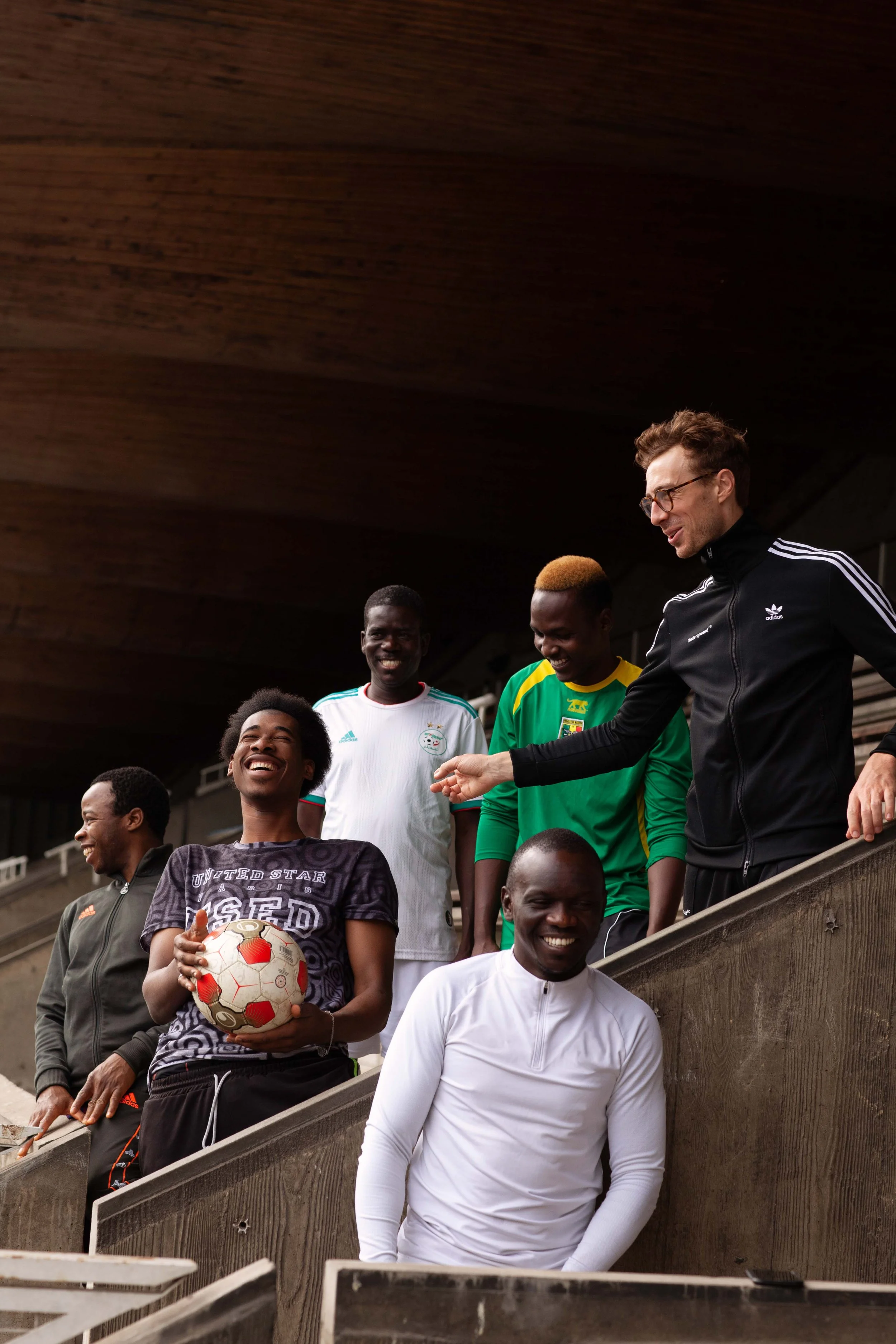 Group of men, including a woman holding a soccer ball, smiling and laughing on stadium seats.