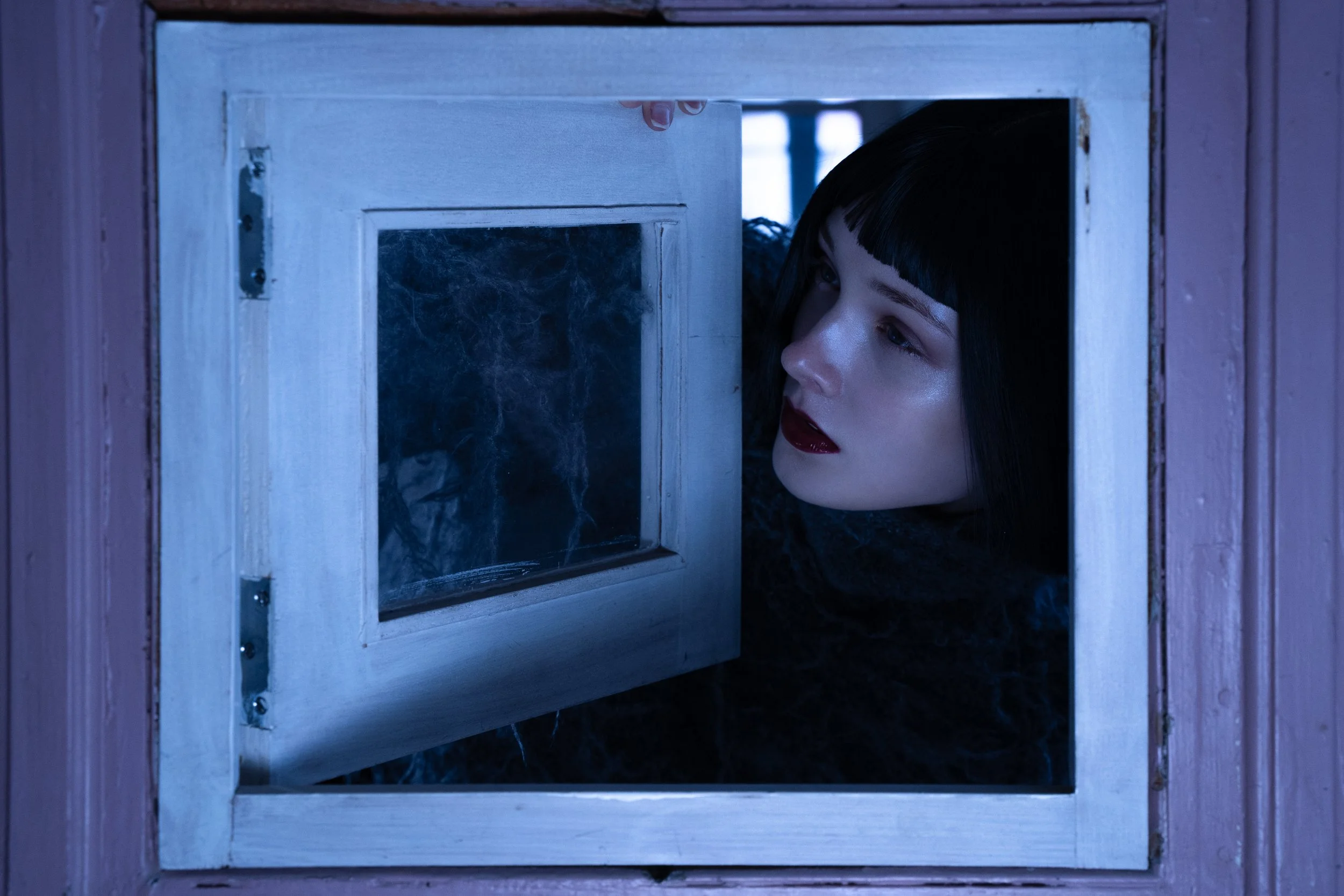 A woman with dark hair and red lipstick looking through a small open window in a pink wall.