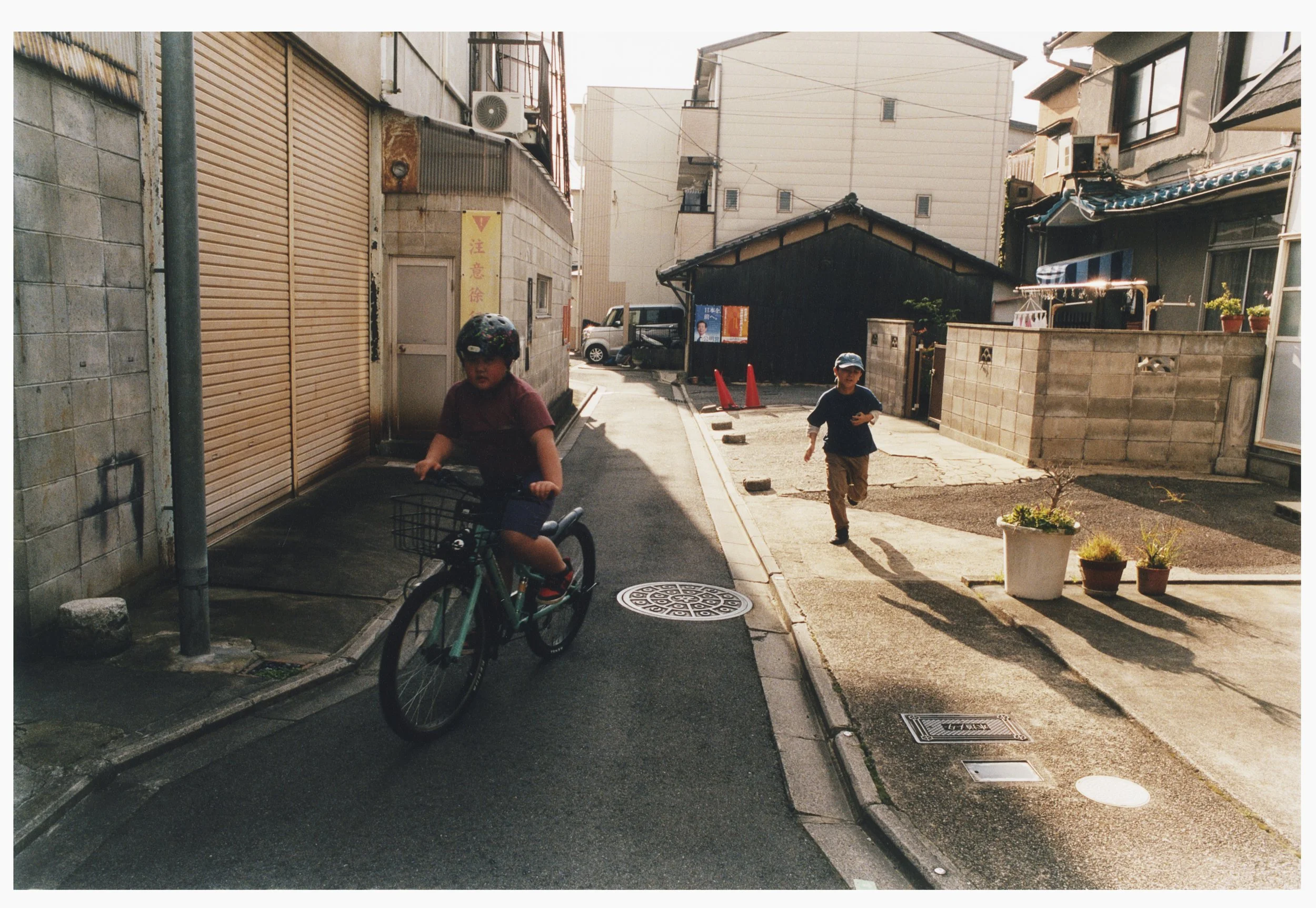 Two children playing on a narrow residential street, one riding a bicycle and the other running. The scene is in a quiet neighborhood with houses and plants, sunlight casting shadows.