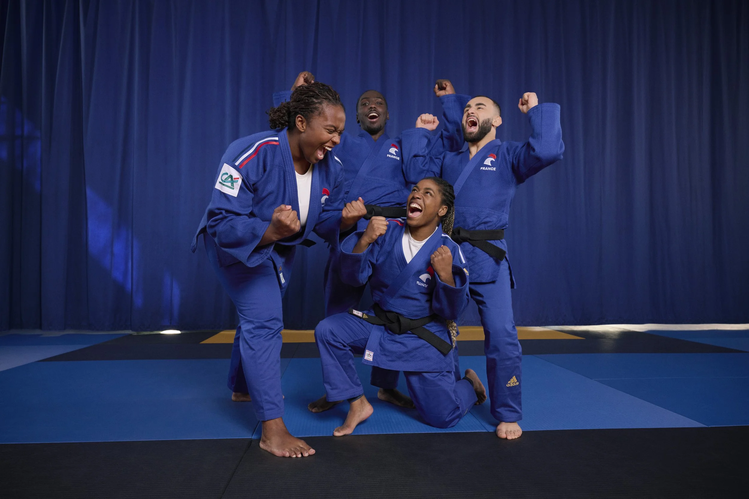 Group of four women and men in judo uniforms celebrating triumph on blue mats with a blue curtain background
