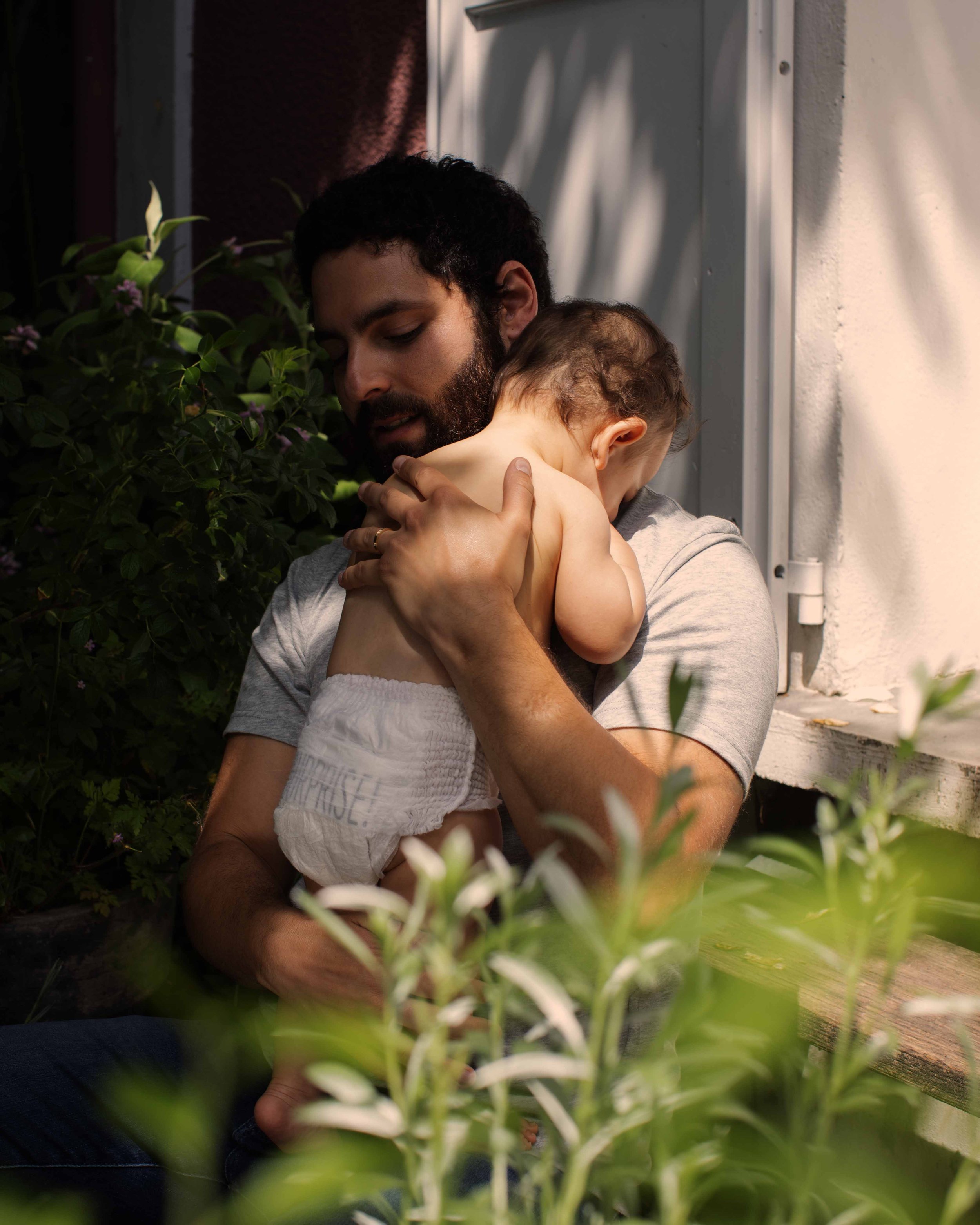 A man holding a young child close to his chest outdoors near a house, with plants and greenery around them.