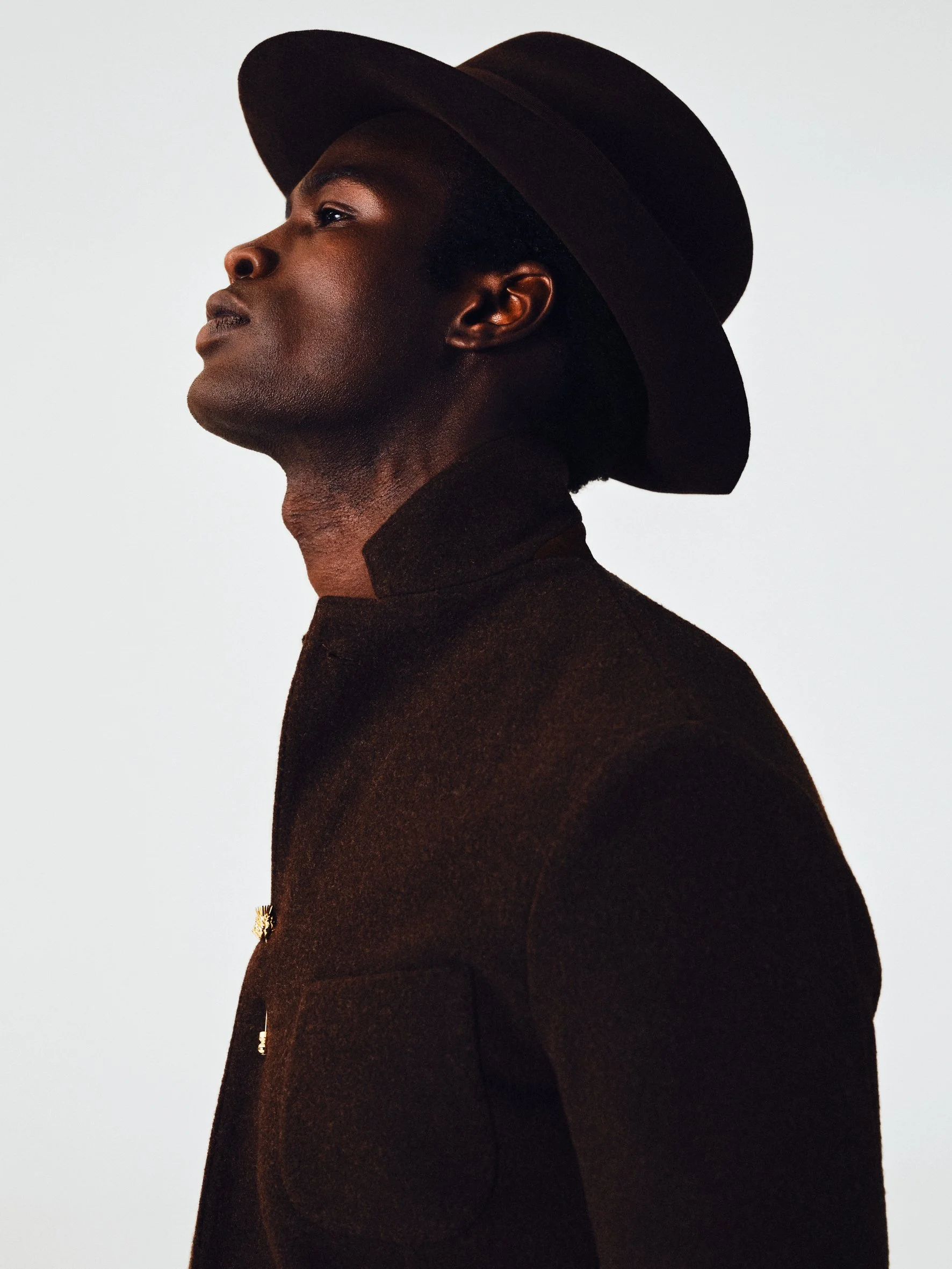A man with dark skin and short curly hair wearing a wide-brimmed black hat and a dark high-collared coat, looking upward and to the side against a plain white background.