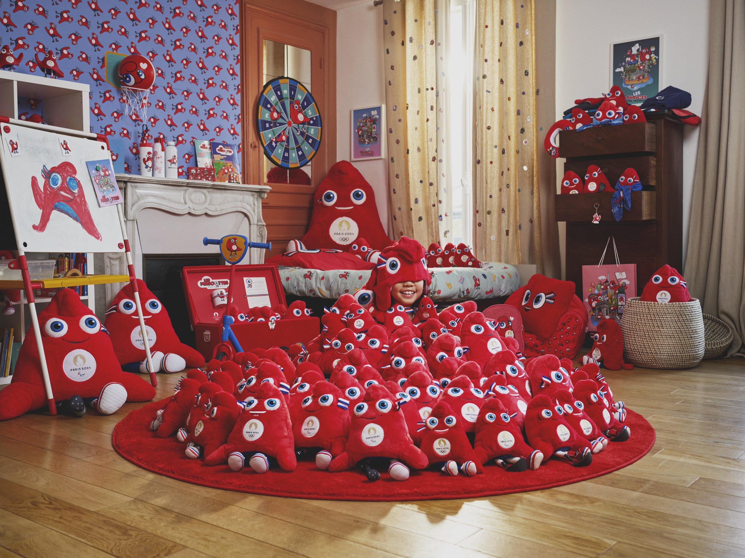 A girl surrounded by numerous red plush toys with big eyes, sitting on a red circular rug in a room decorated with red, blue, and white colors, featuring a white fireplace, artwork, and toys related to the Paris 2024 Olympics.