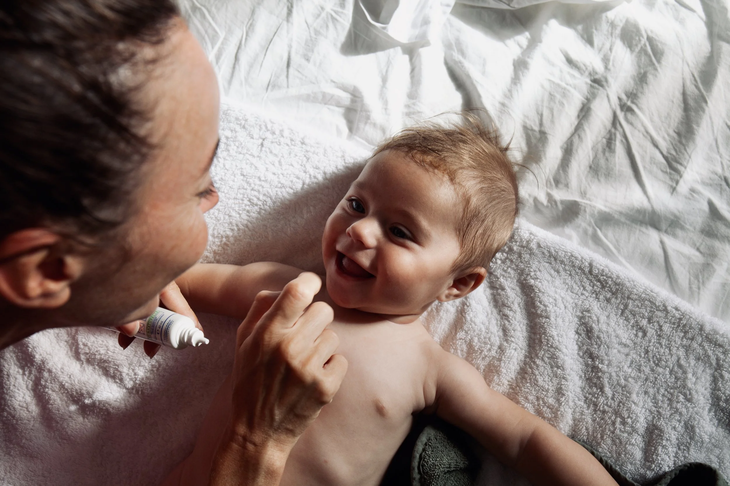 Adult applying ointment or lotion to smiling baby lying on a white towel on a bed.