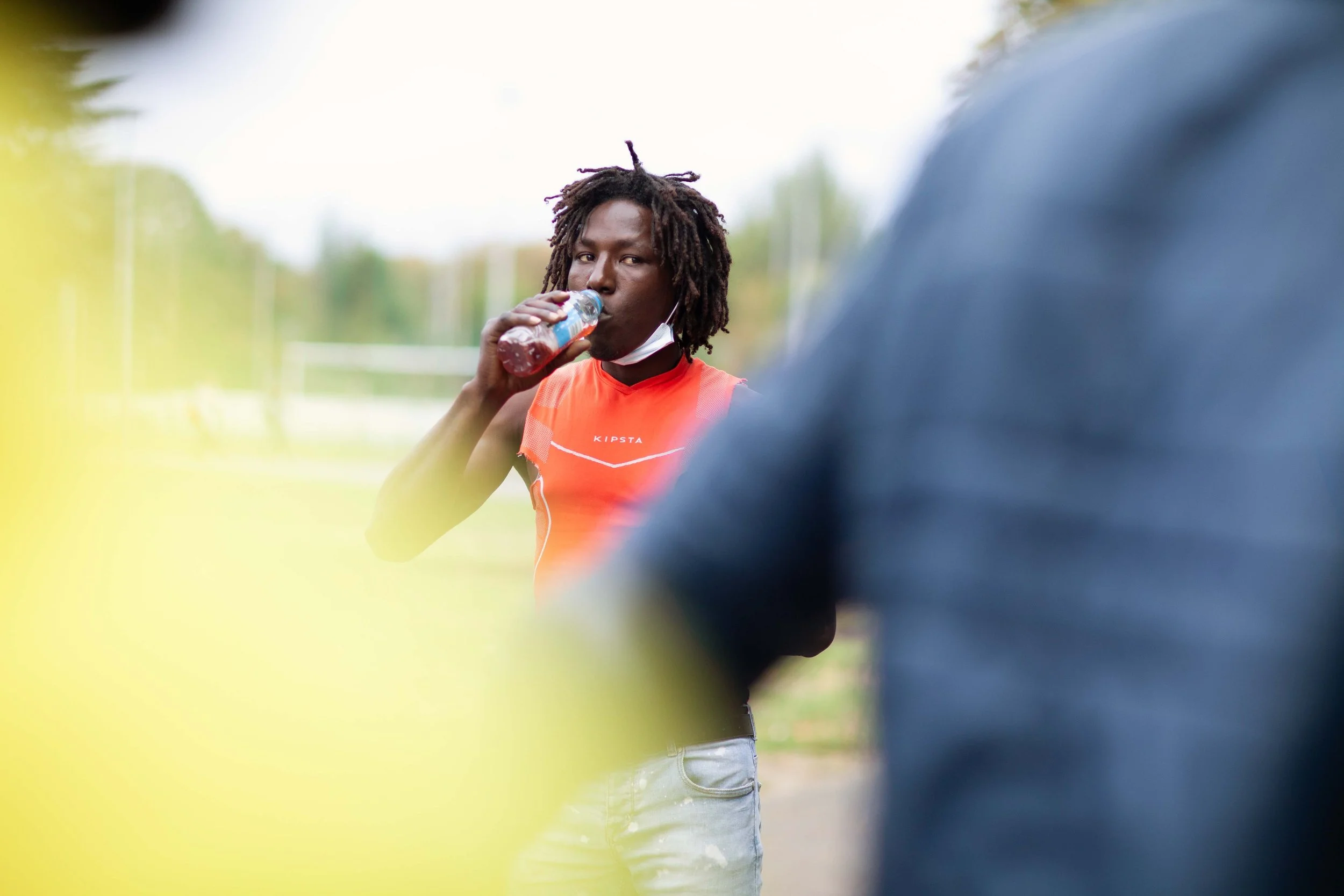 A young man with dreadlocks, wearing an orange sports jersey, is drinking from a water bottle. There are blurred figures and trees in the background.