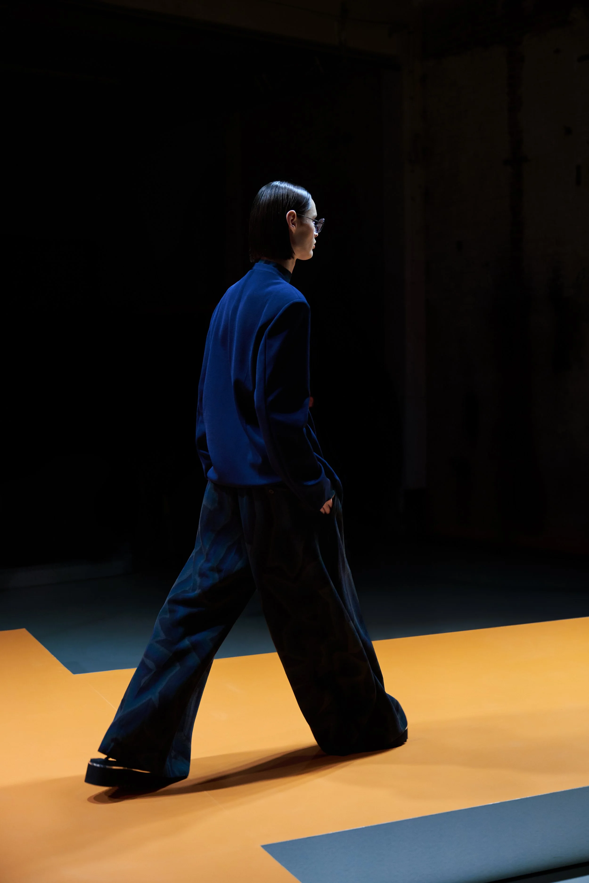 A woman with short dark hair and glasses walking on an orange floor in a dark room, wearing a blue blazer and loose dark pants.