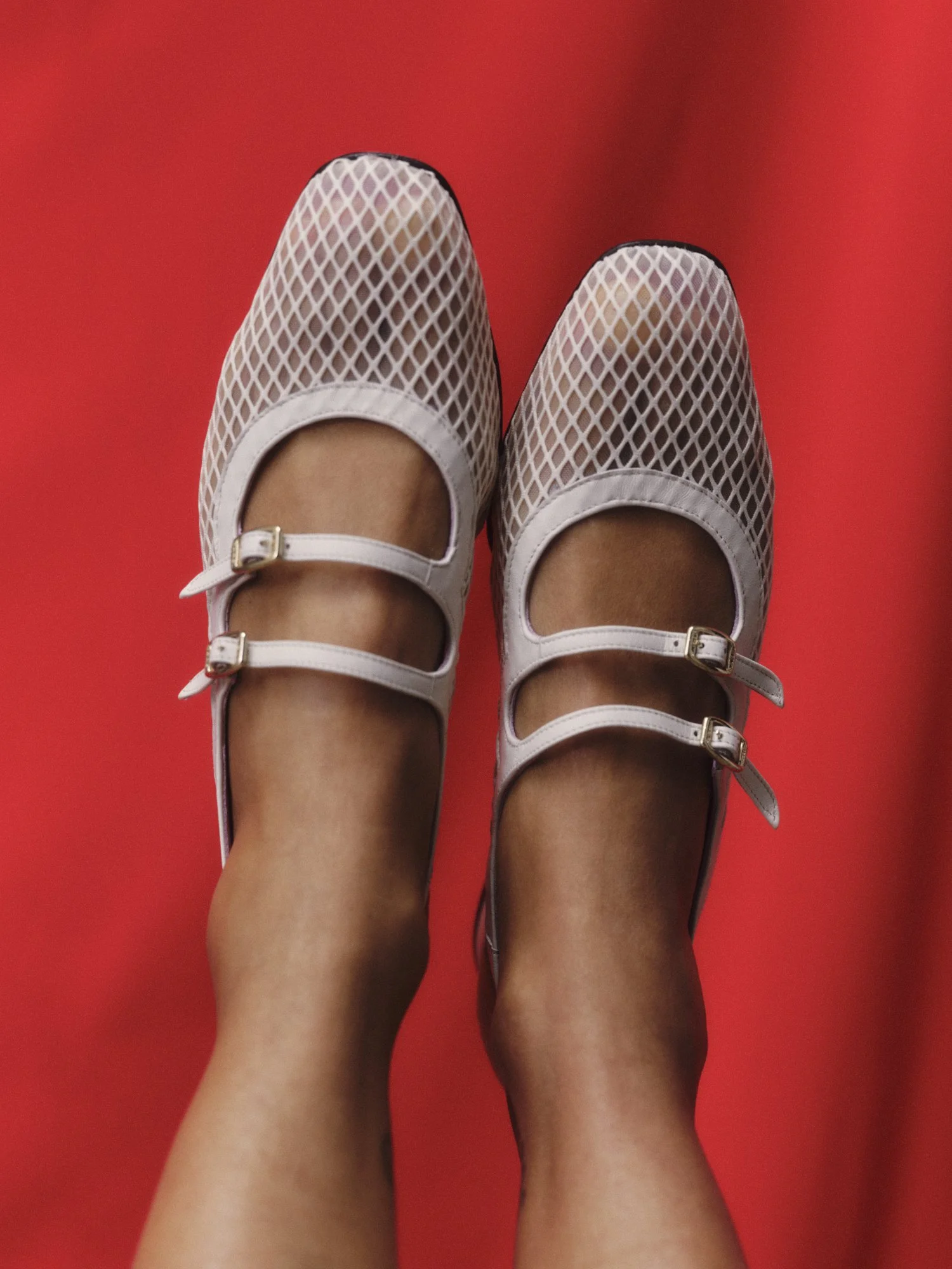Two feet wearing white fishnet shoes with ankle straps over a red background.