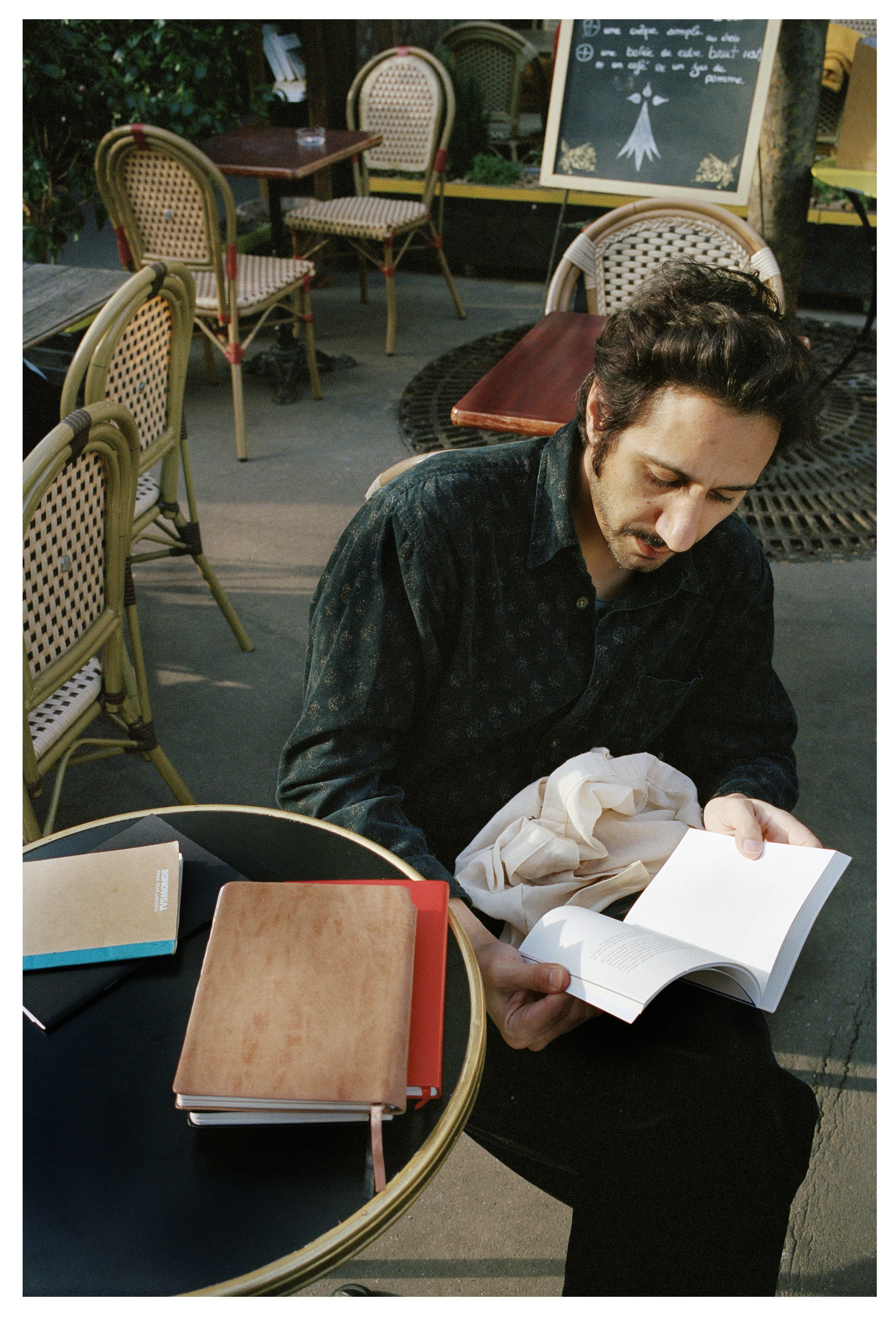A man sitting at an outdoor cafe, reading a book, with several notebooks and a pen on the table in front of him.