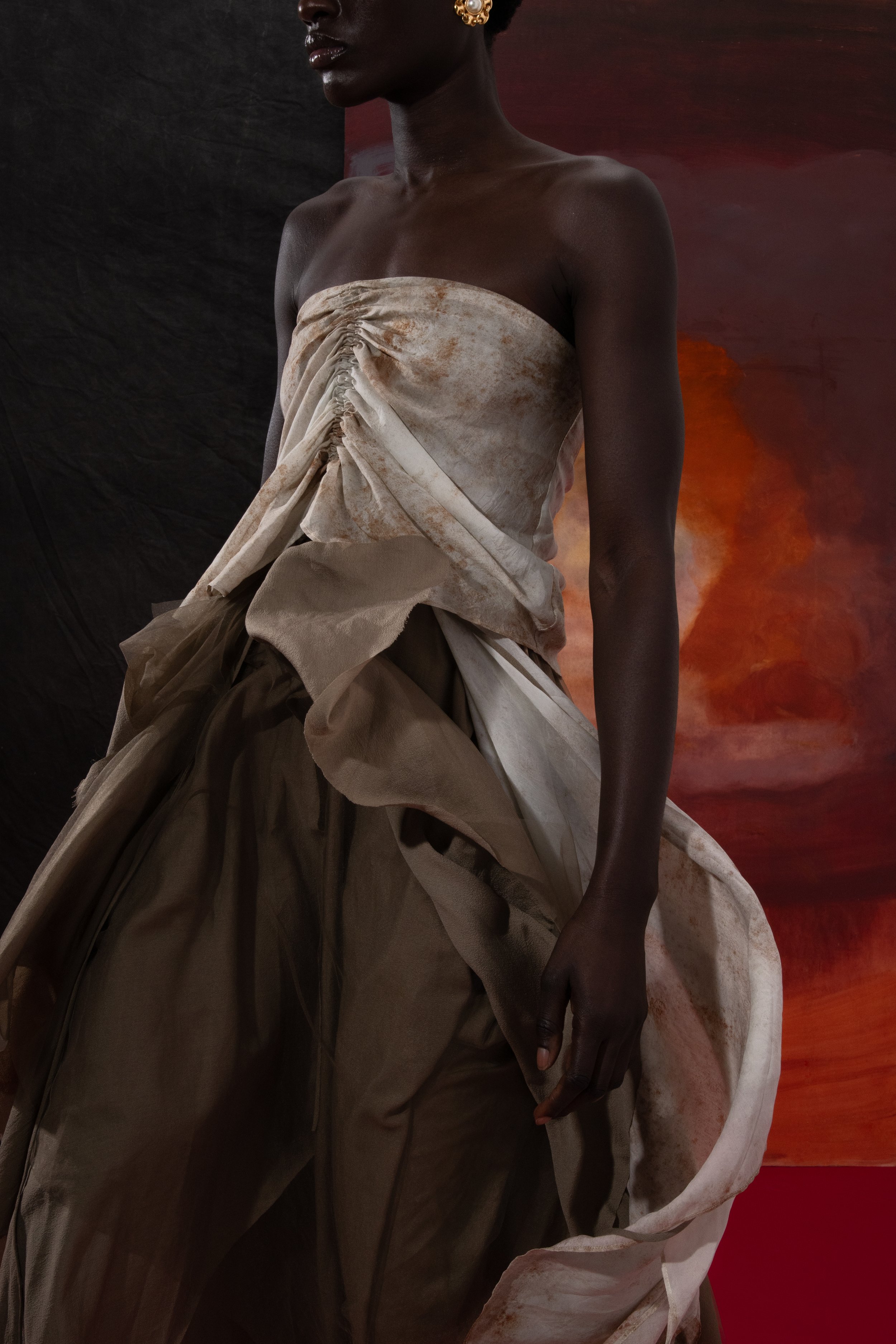 A woman with dark skin wearing an elegant, beige, and champagne-colored dress with ruching and draping, standing against a dark background with warm reddish tones.