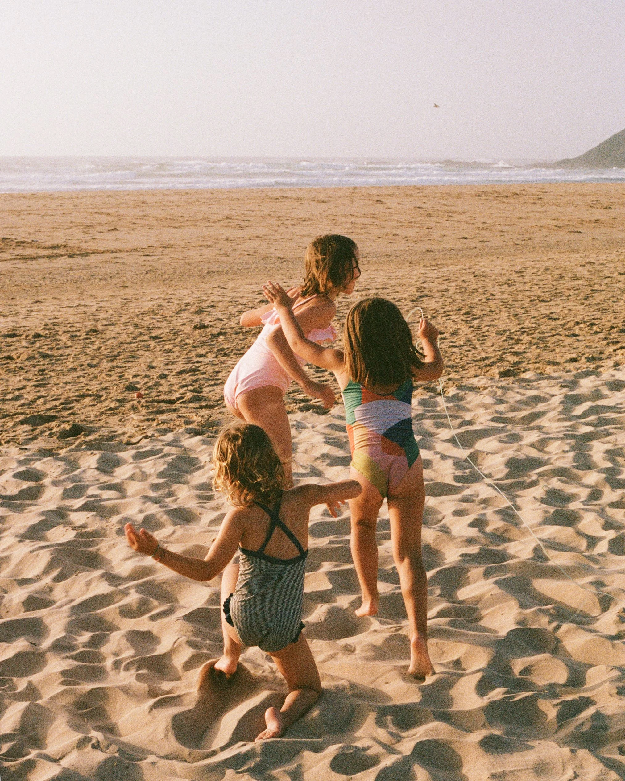 Three children playing in the sand on a beach with the ocean in the background, two girls and a younger girl, one girl holding a string, possibly a kite, during sunset.