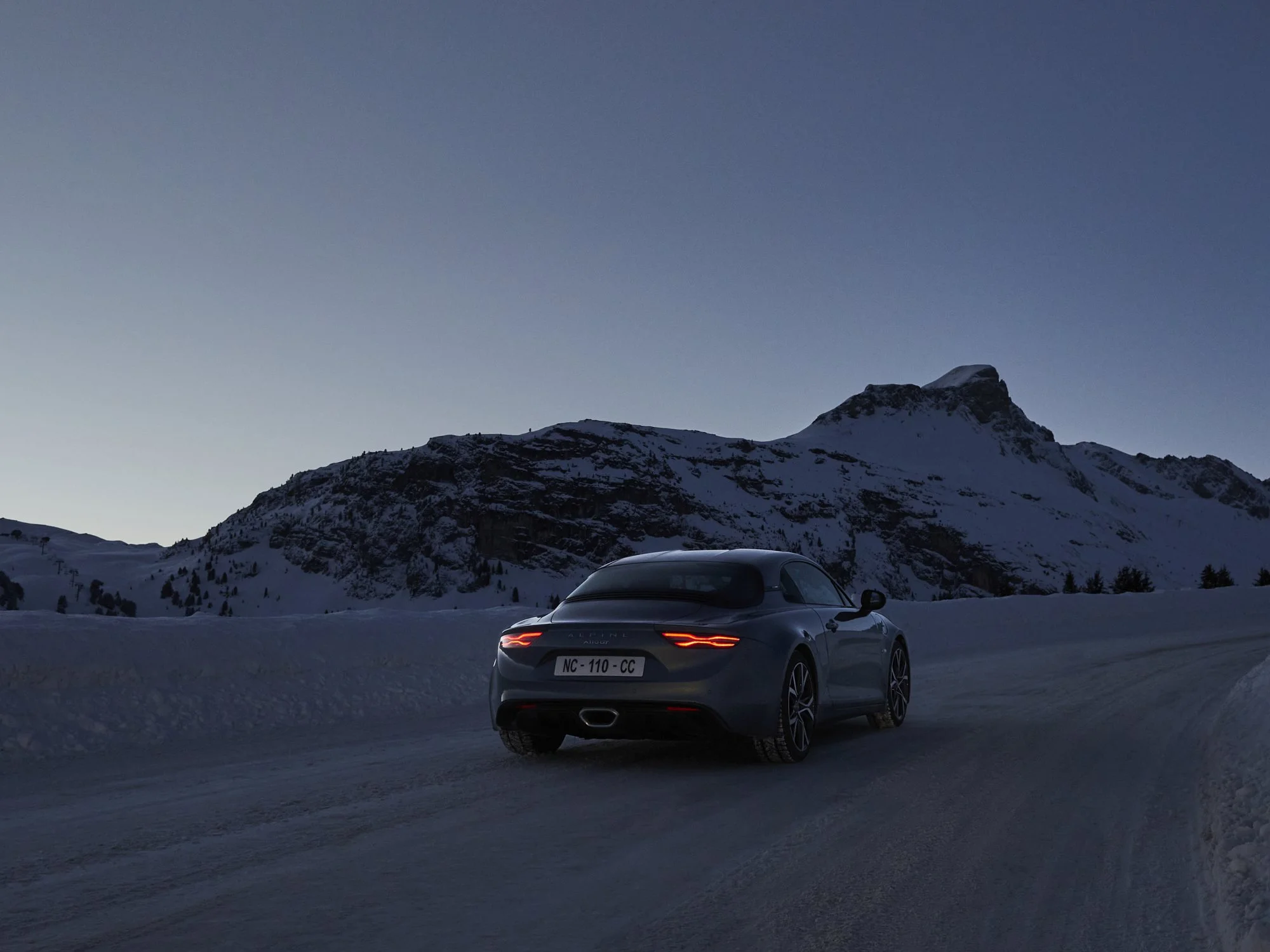 A dark gray luxury convertible car driving on an icy mountain road at dusk with snow-covered mountains and a clear sky in the background.