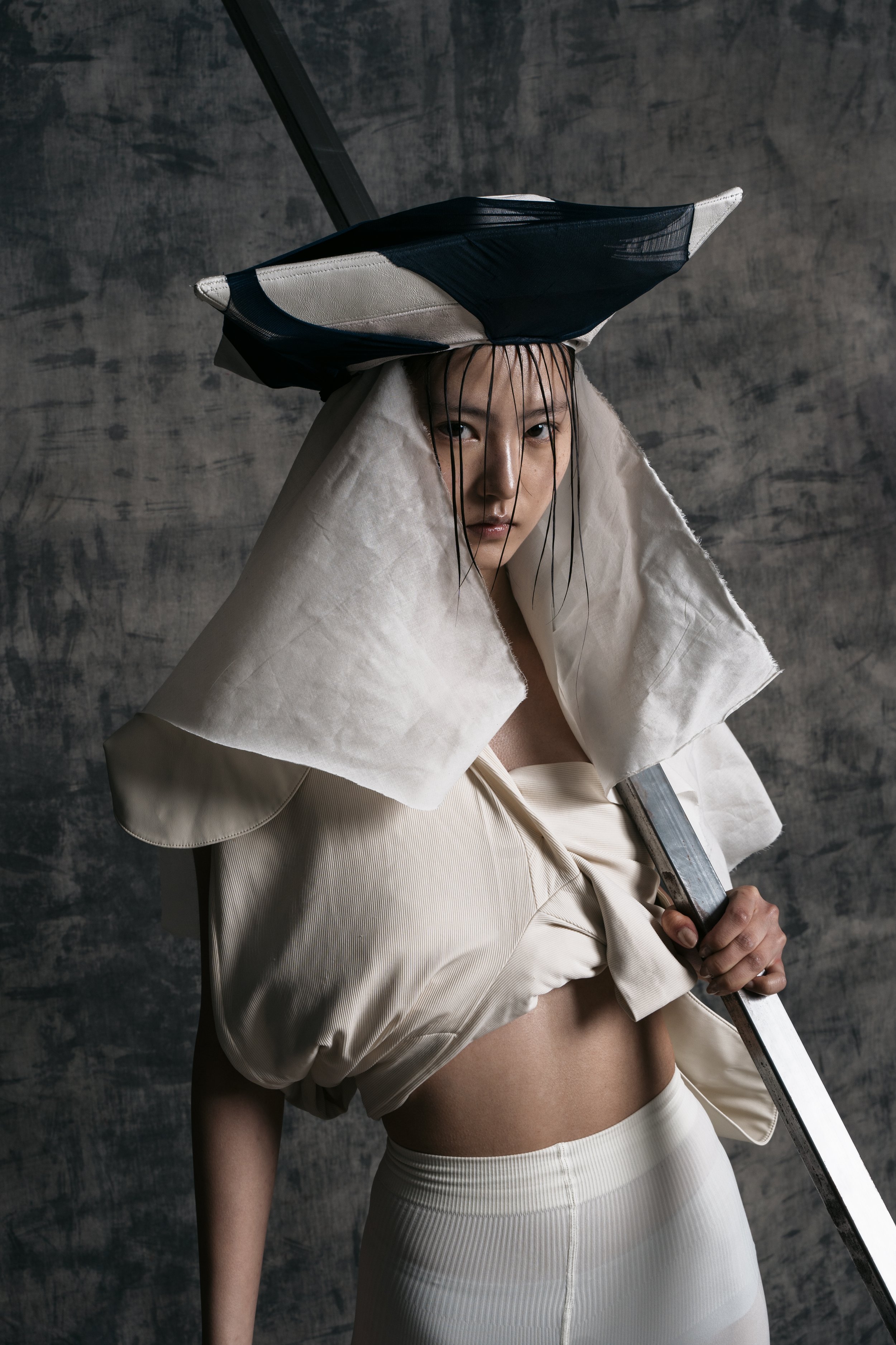 A woman with wet hair, dressed in cream-colored clothing, holding a large sword. She wears an oversized cream coat and a wide-brimmed black and white hat. The background is a textured gray wall.