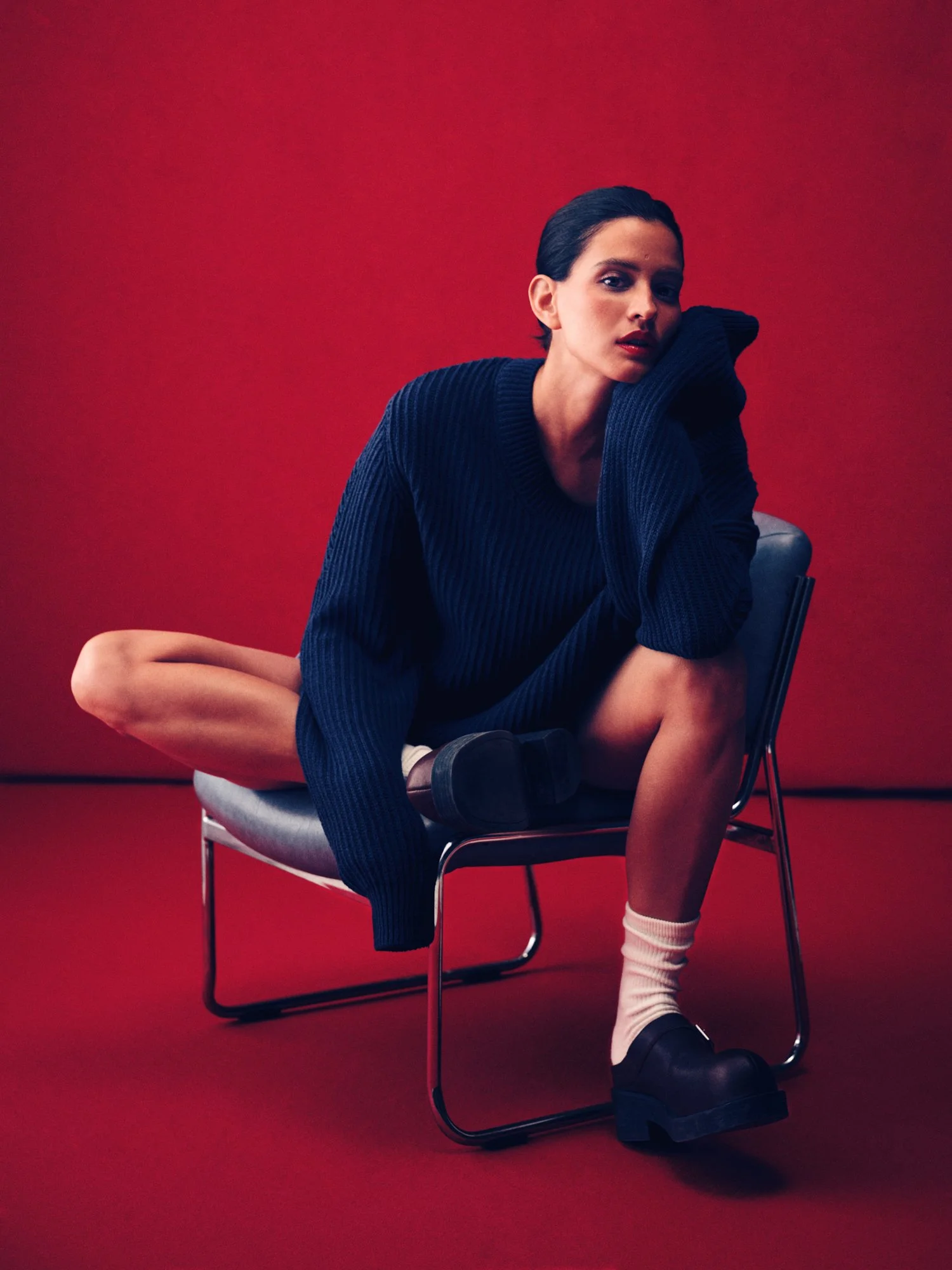 A woman with dark hair in a navy oversized sweater, sitting on a modern chair with one leg crossed over the other against a red background.