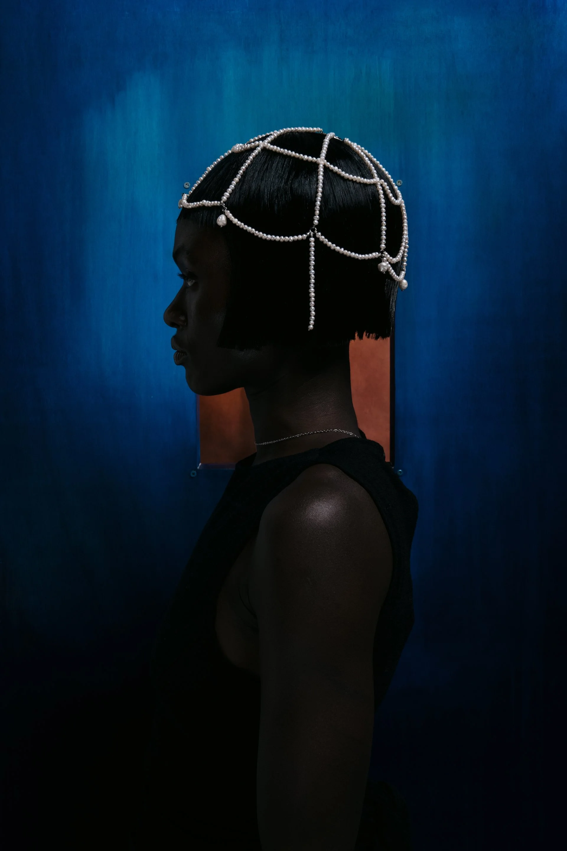 Profile of a woman with short black hair wearing pearl jewelry, standing against an abstract background.