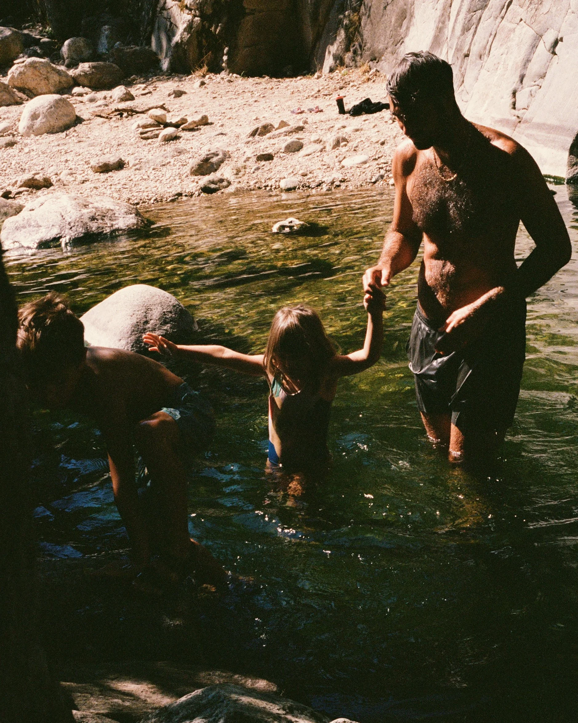 A man and two children enjoying a swim in a natural creek surrounded by rocks and cliffs, with one child helping another.