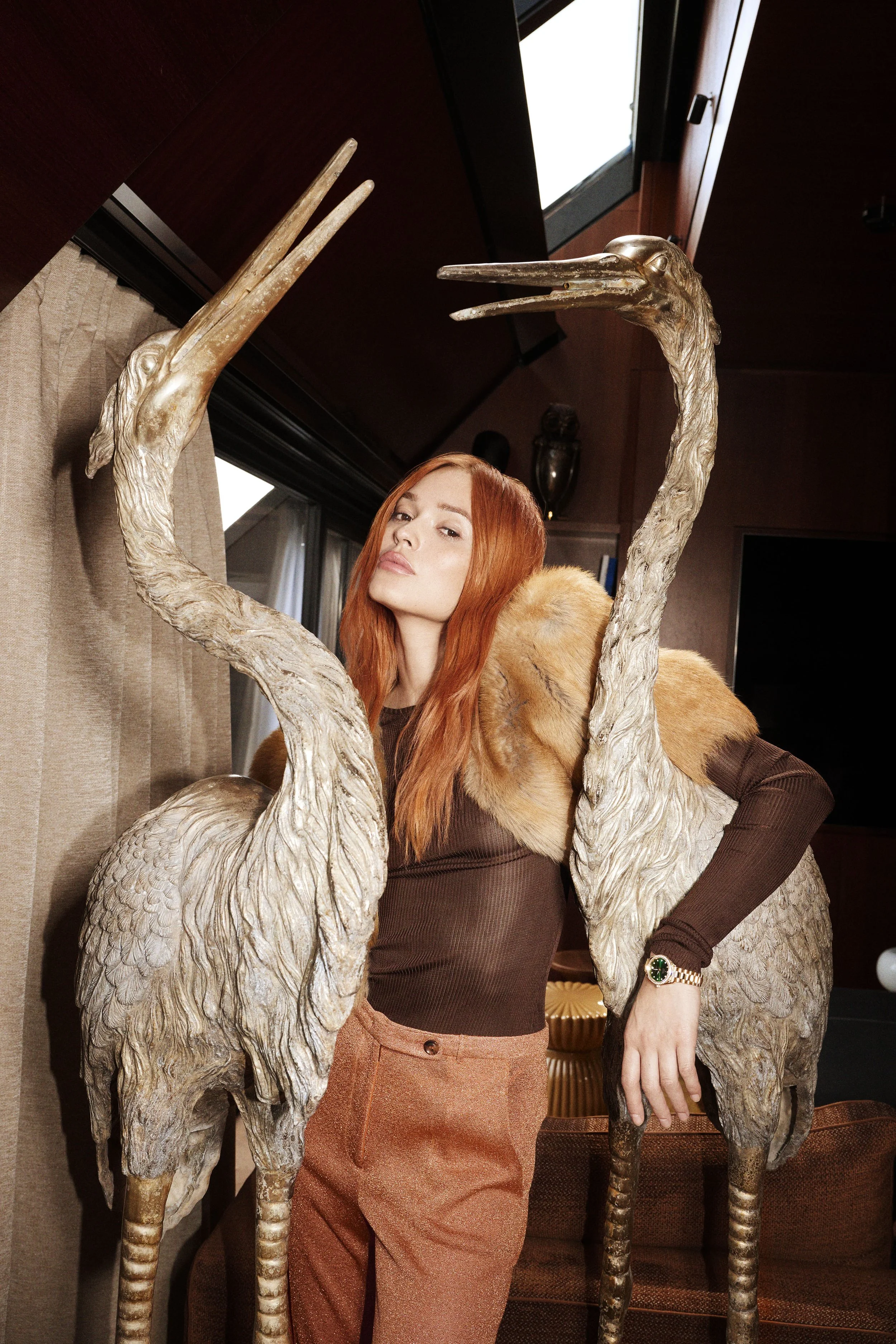 A woman with long red hair and a brown outfit standing between two large decorative American white-stack herons with long necks and beaks in an indoor setting.