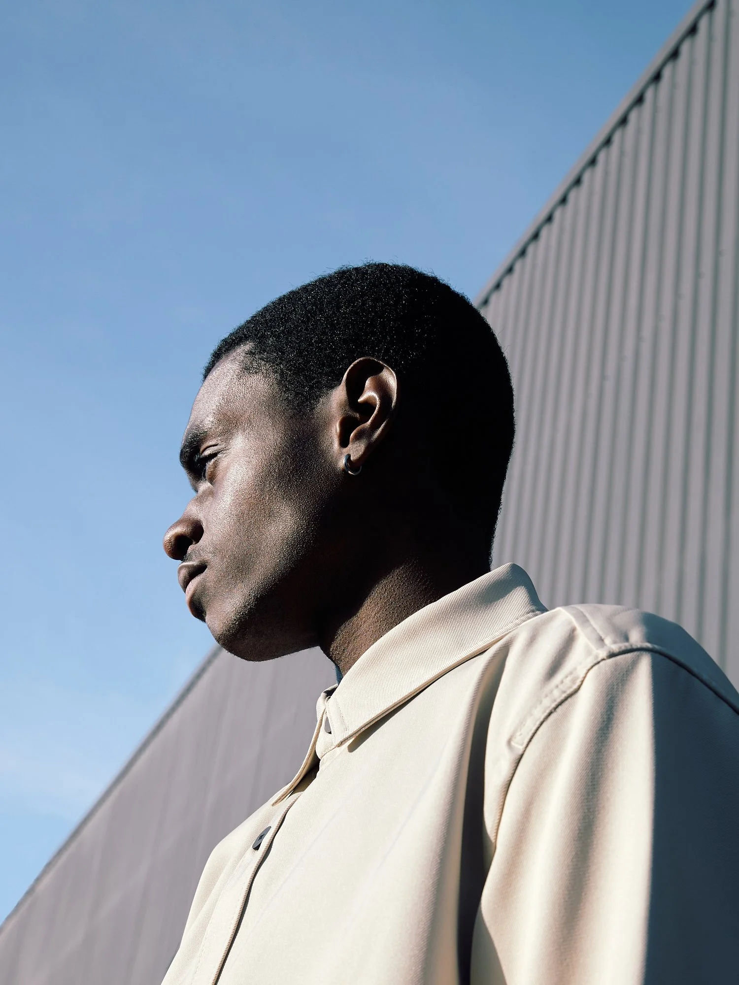 Side profile of a young man with a short hairstyle, wearing a beige jacket, standing outdoors near a metal building with a clear blue sky in the background.