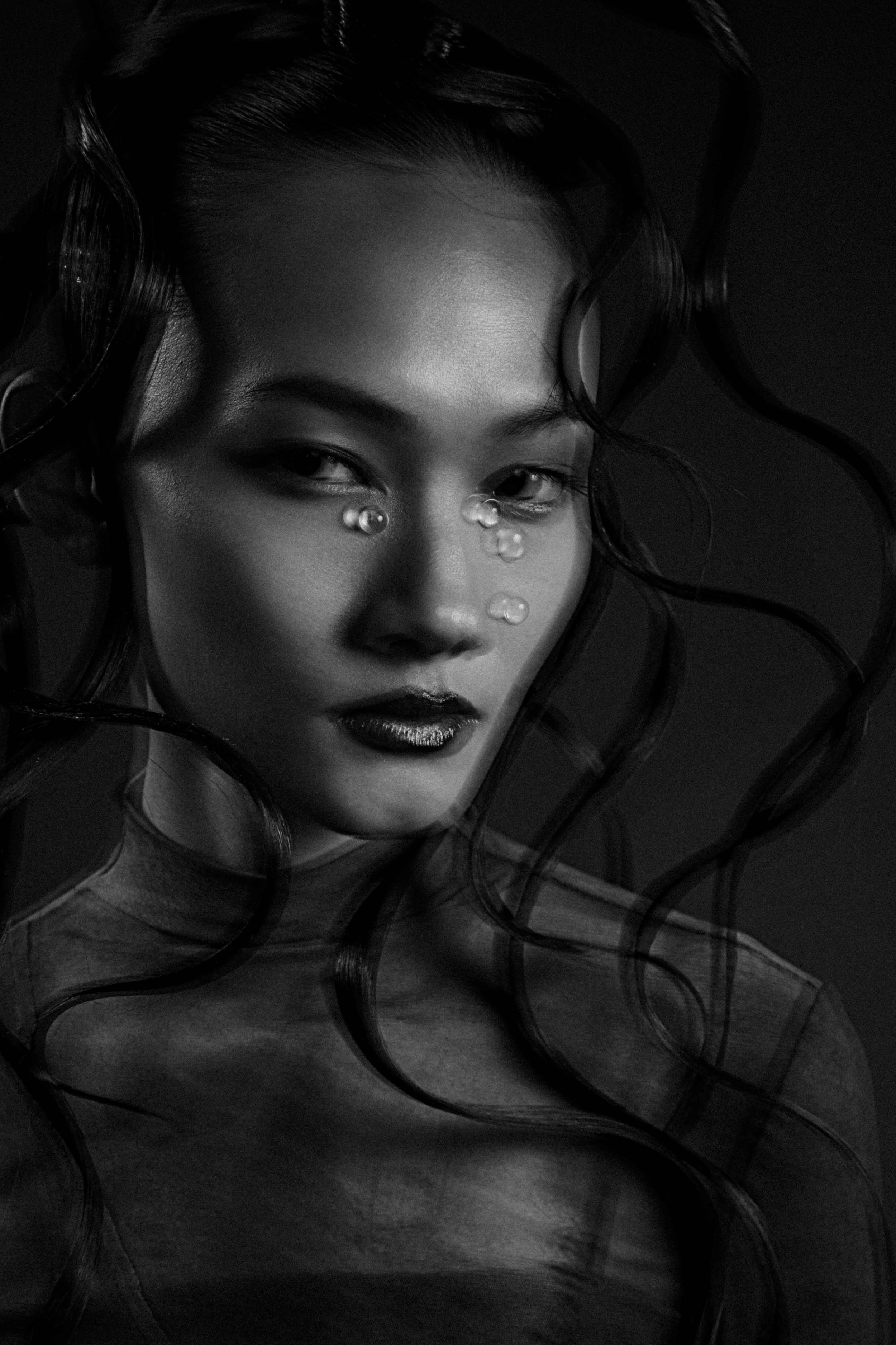 Black and white portrait of a woman with wavy hair, shimmering makeup, and facial pearls, looking confidently at the camera.