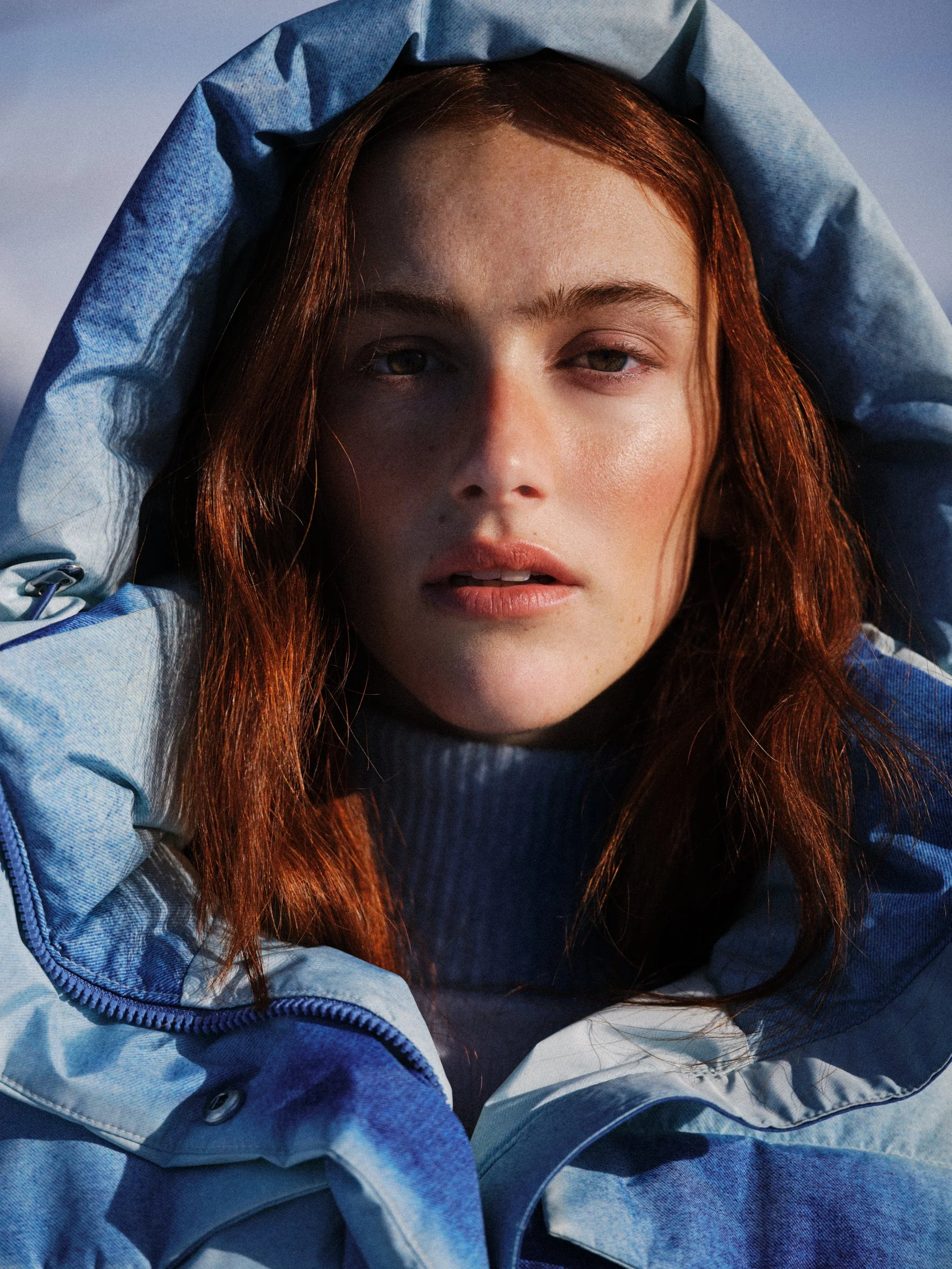 A young woman with red hair looking directly at the camera, wearing a blue hooded jacket, outdoors during what appears to be late afternoon or early evening.