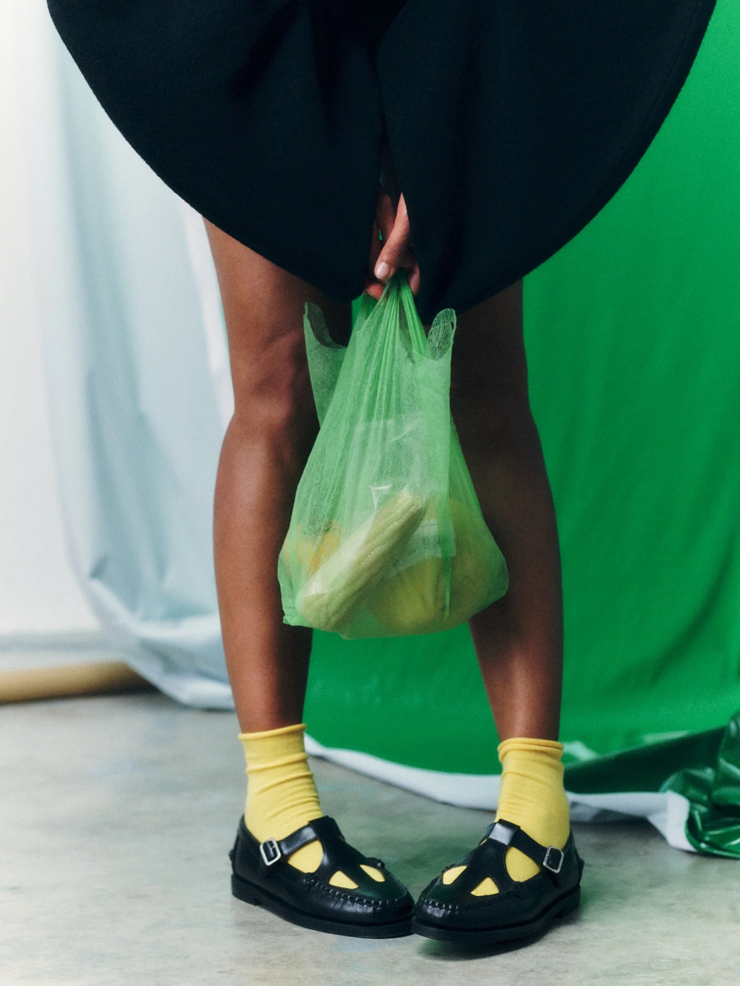 Close-up of a person holding a green plastic bag filled with bananas, wearing black shoes with buckles, yellow socks, a black skirt, and yellow wristbands, standing in front of a green background.