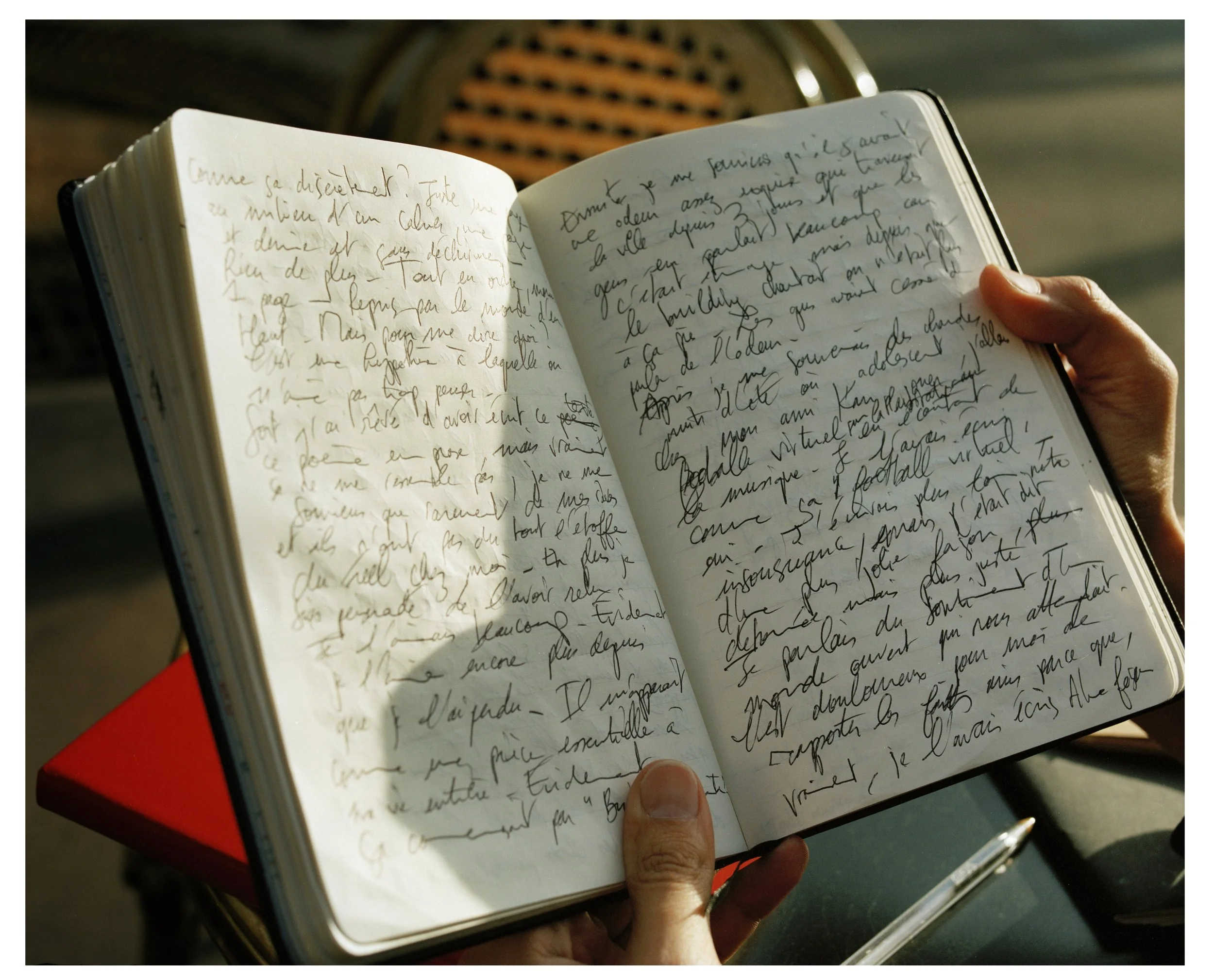 A person holding open a handwritten journal with French cursive writing, with a red book underneath, and a pen resting nearby. The background shows a washing machine drum.