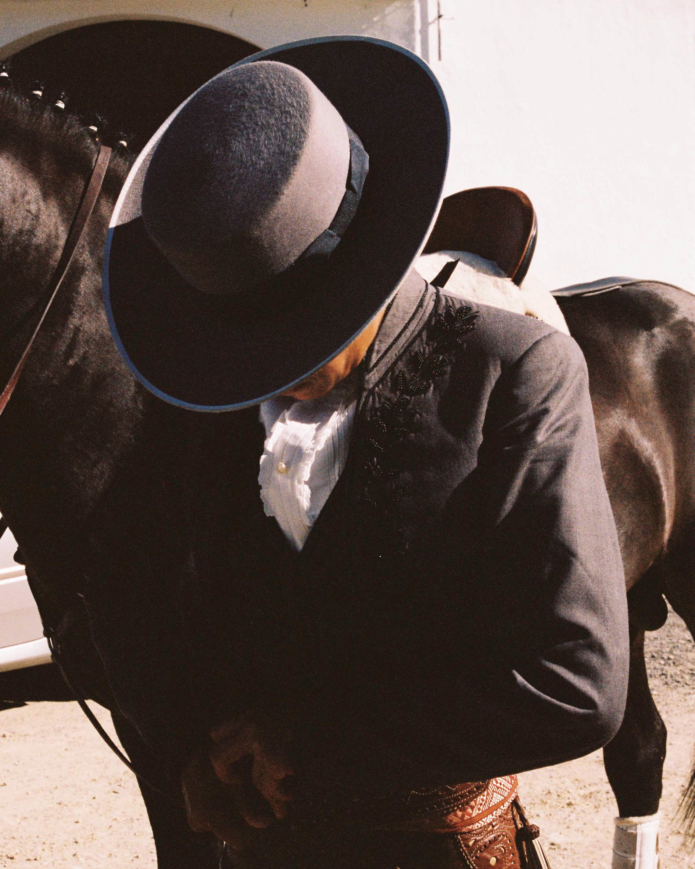 A person wearing a large hat, a dark blazer, and patterned pants standing next to a horse.