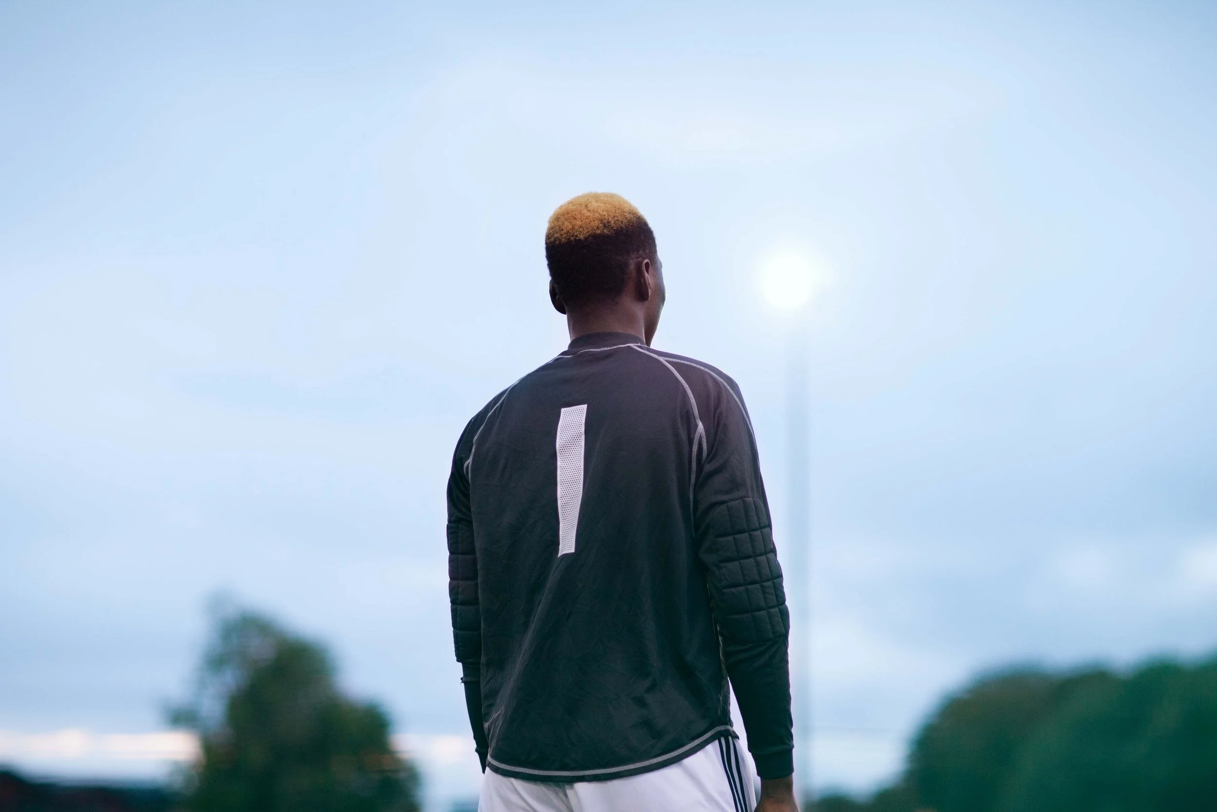 A person with blonde hair, wearing a black sports jacket with the number 1 on the back, standing on a field at dusk or dawn, facing away from the camera, with trees and a cloudy sky in the background.