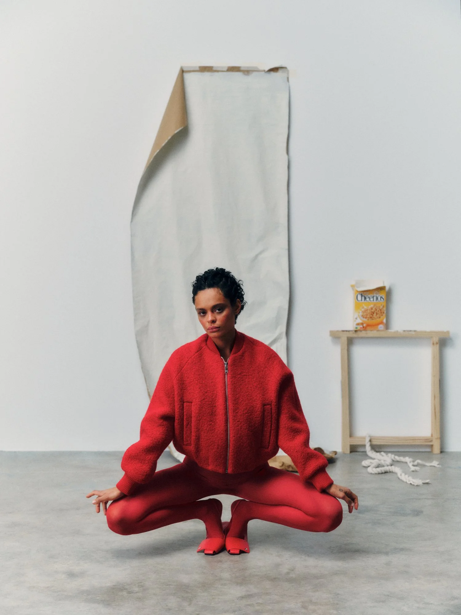 A woman in a red jacket and matching red tights is sitting cross-legged on the floor in front of a plain wall. Behind her, there is a tall mirror, a small wooden shelf with a box of Cheerios cereal, and a coiled rope on the floor.