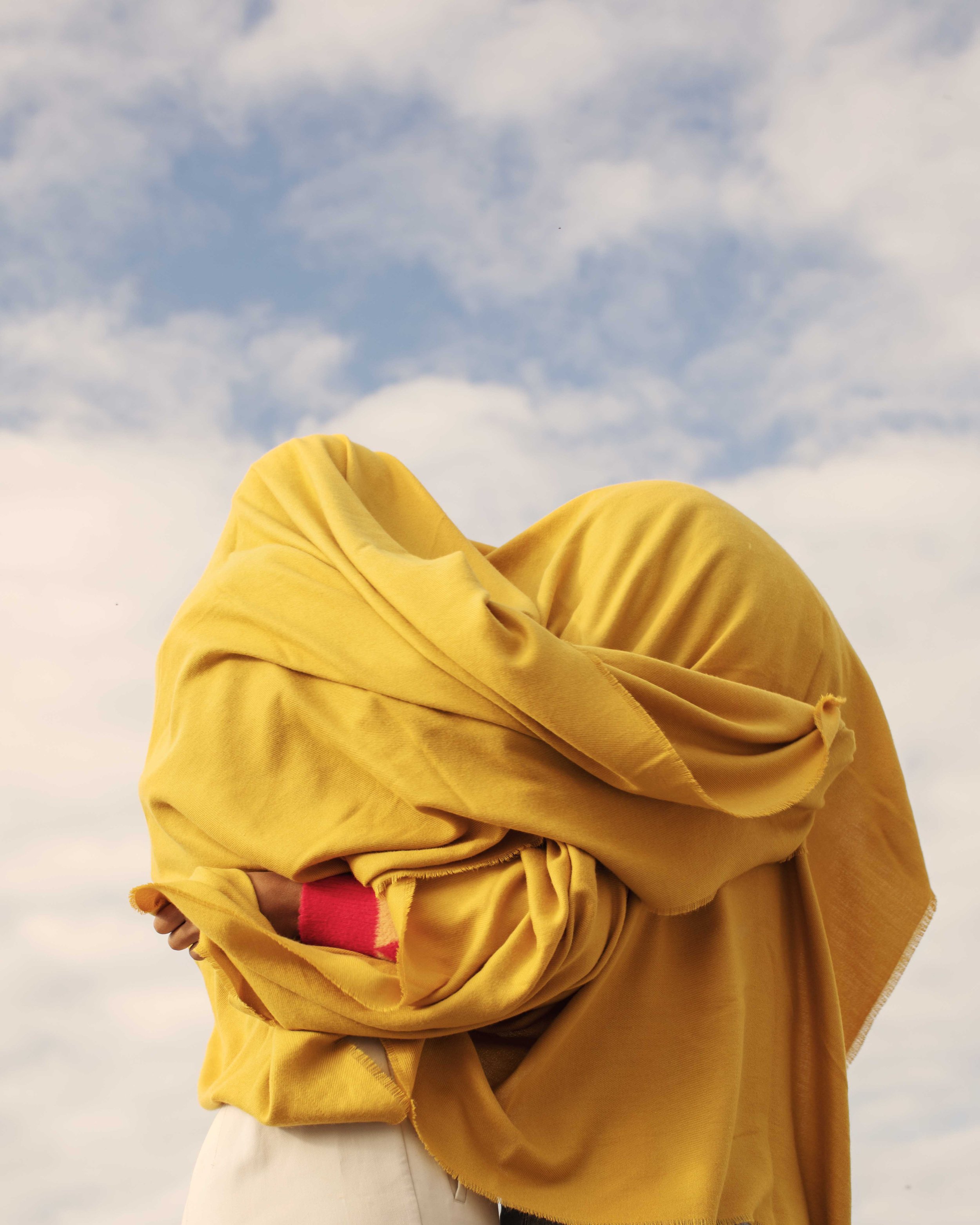 Person wearing a yellow hoodie and pink shirt holding their arm with a cloudy sky in the background.
