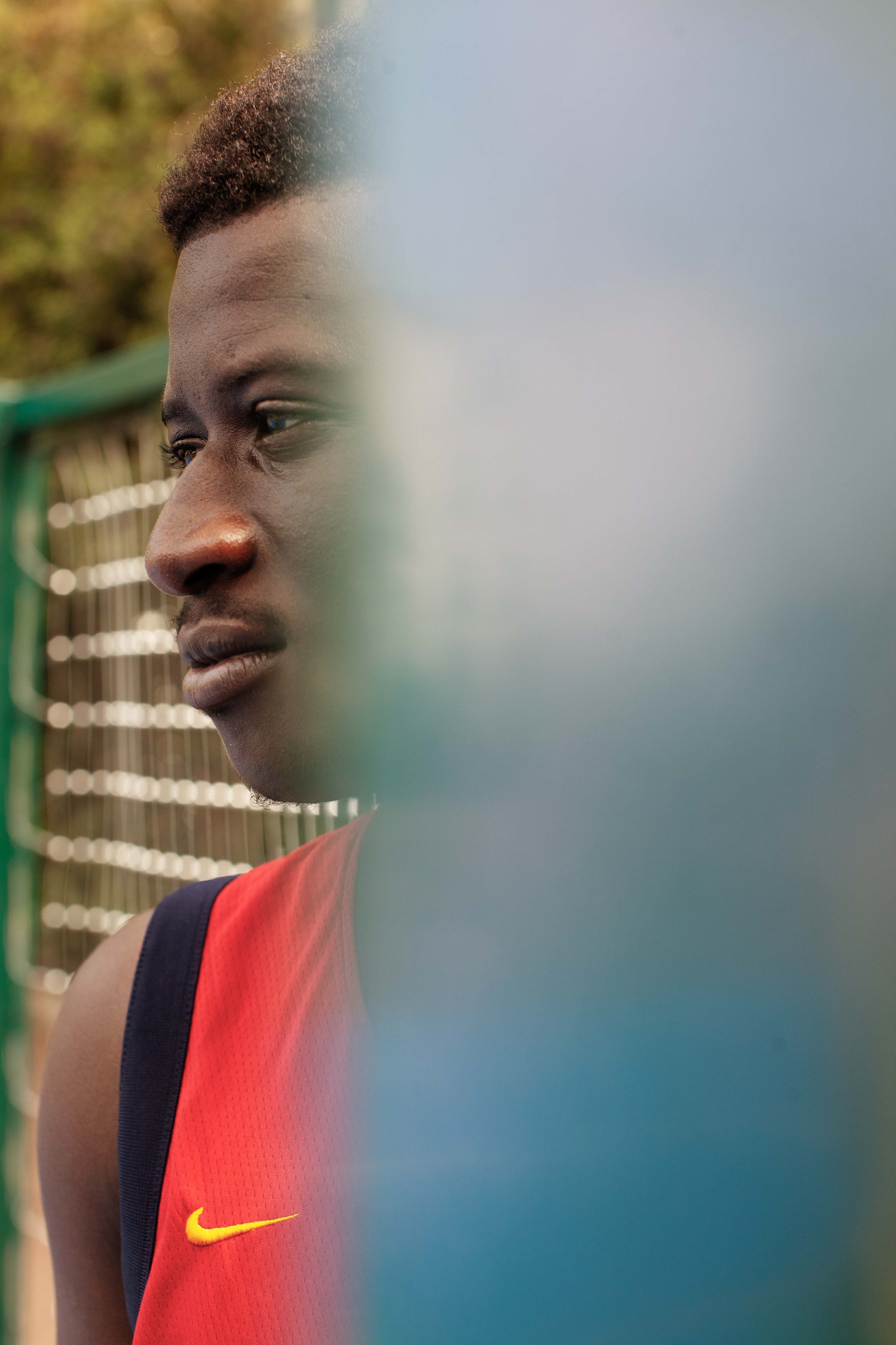 Close-up of a young man with short hair wearing a red sports jersey with a yellow Nike logo, sitting outdoors near a green fence, looking pensively to the left, with trees in the background.
