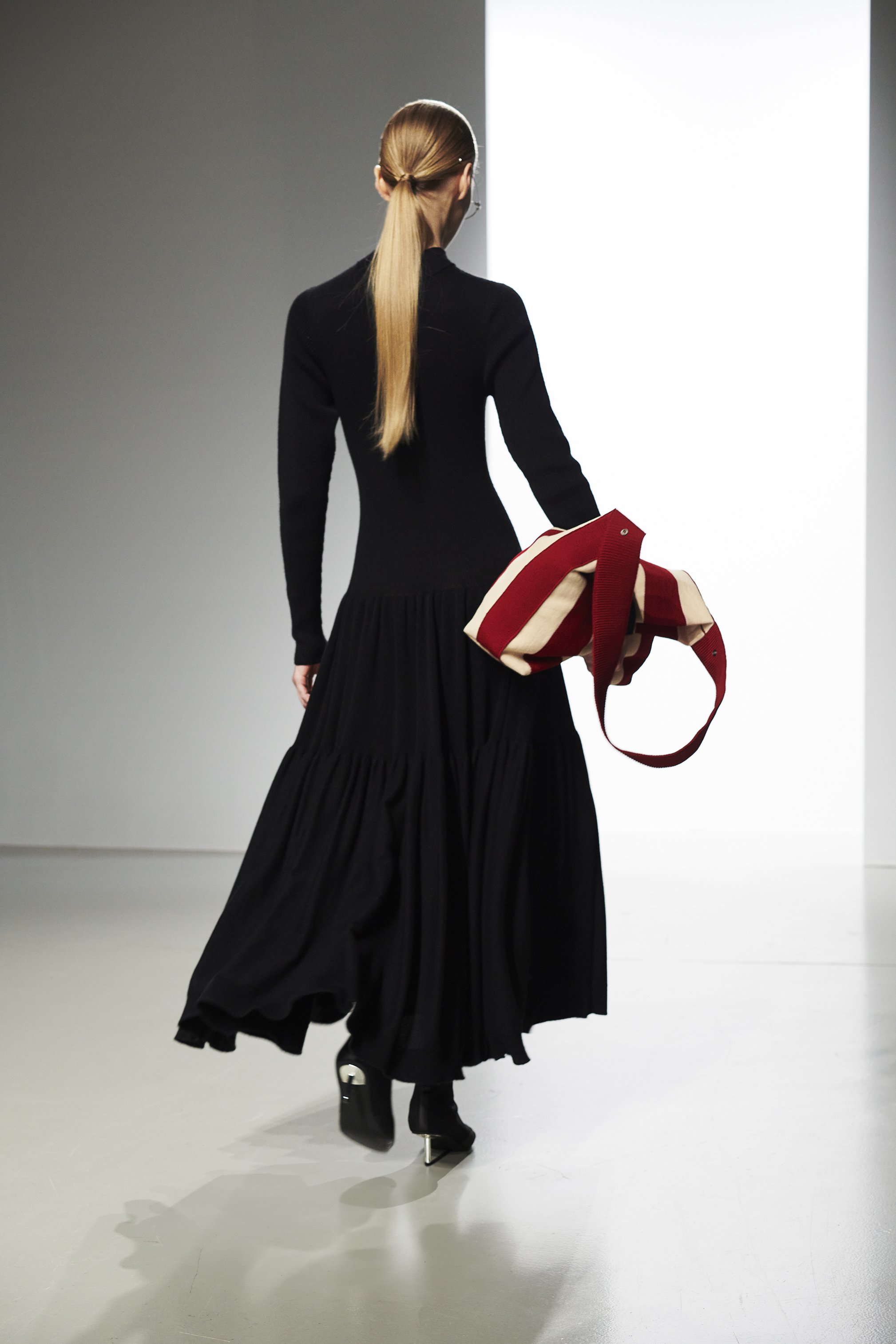 A woman with long blonde hair walking in an art gallery, wearing a long black dress and uphill boots, holding a red and beige striped bag.