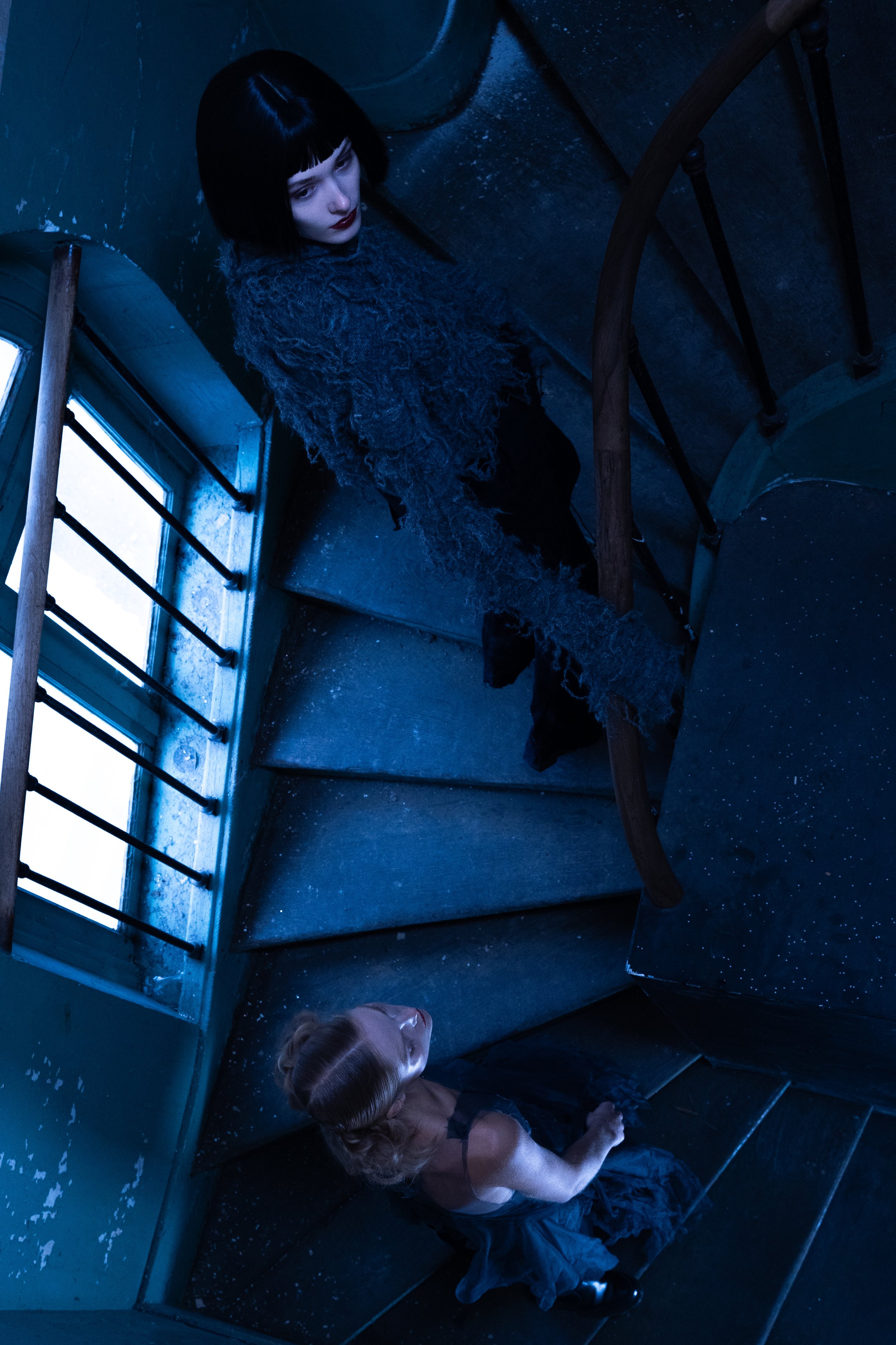 A woman with dark hair and makeup standing on a staircase looking down at a blonde woman sitting on the floor, both in a dark, blue-lit setting.