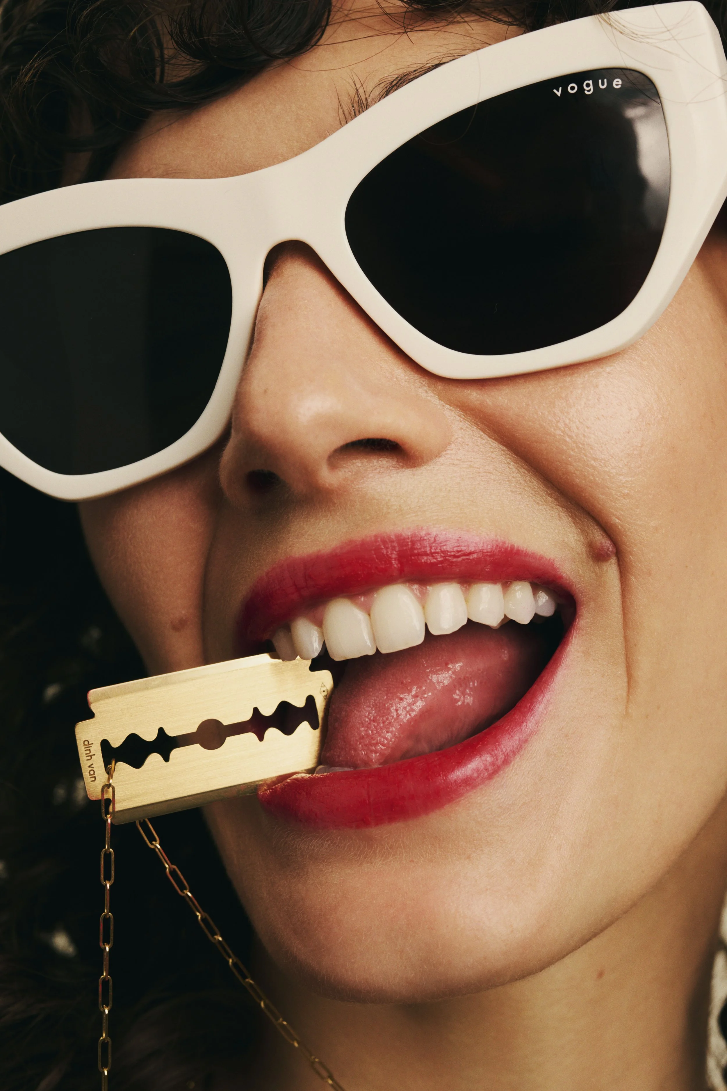 A woman with red lipstick wearing large white sunglasses smiling, with a rare straight razor blade in her mouth.