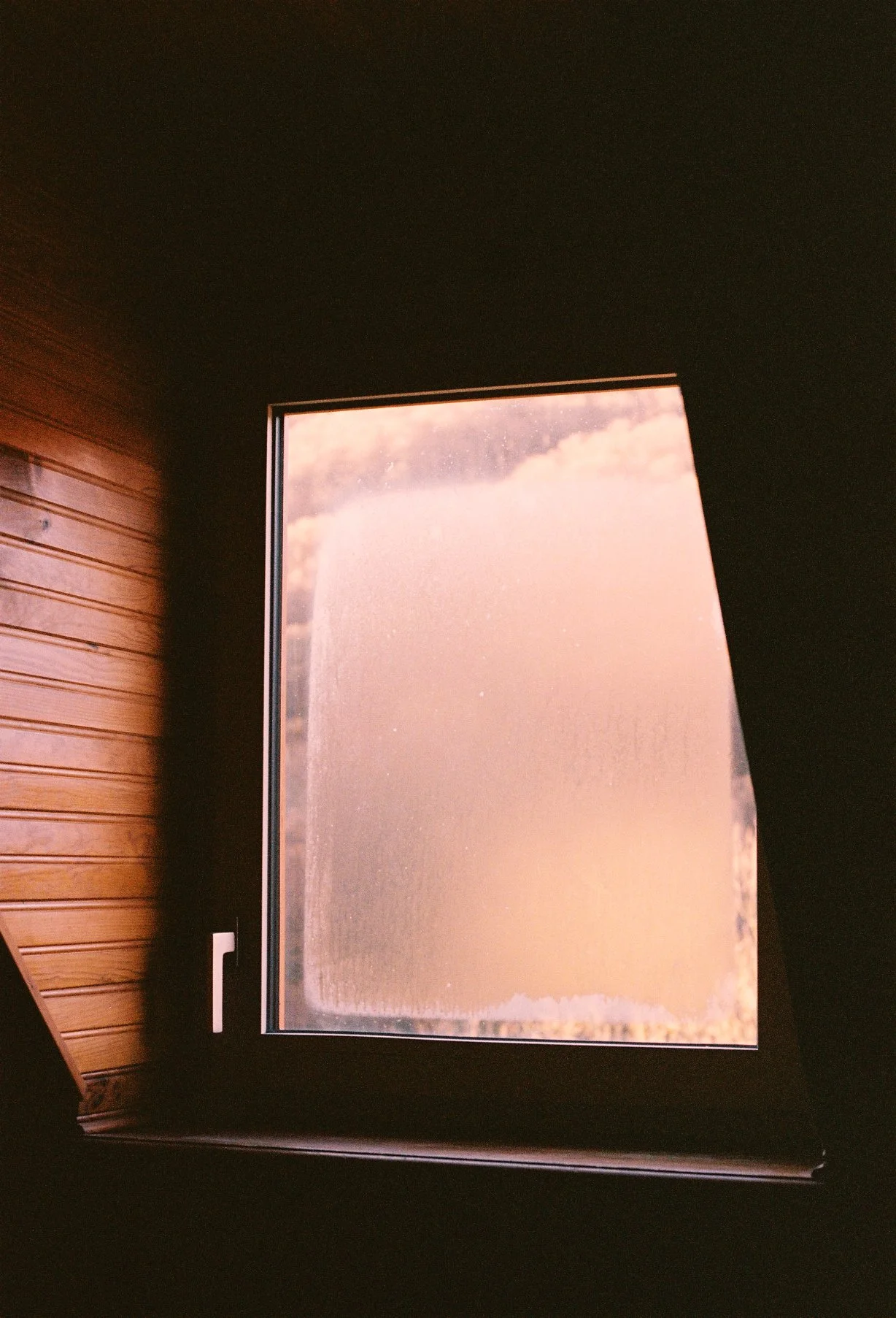 A window with a view of clouds and sky, surrounded by a dark interior wall and a wooden wall on the left side.