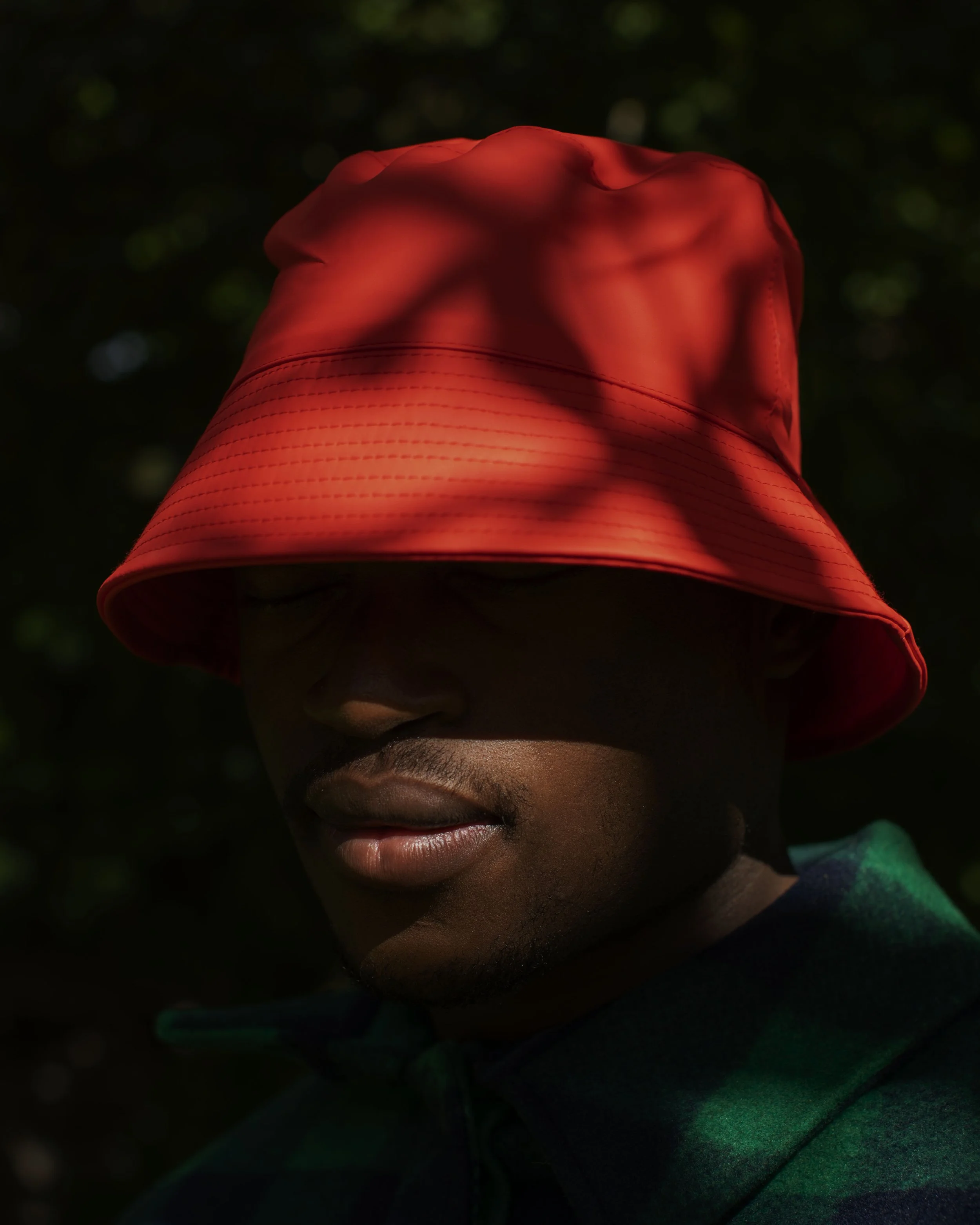Person wearing an orange bucket hat covering their eyes, with visible lips and nose, wearing a green and black jacket, in a dark outdoor setting.