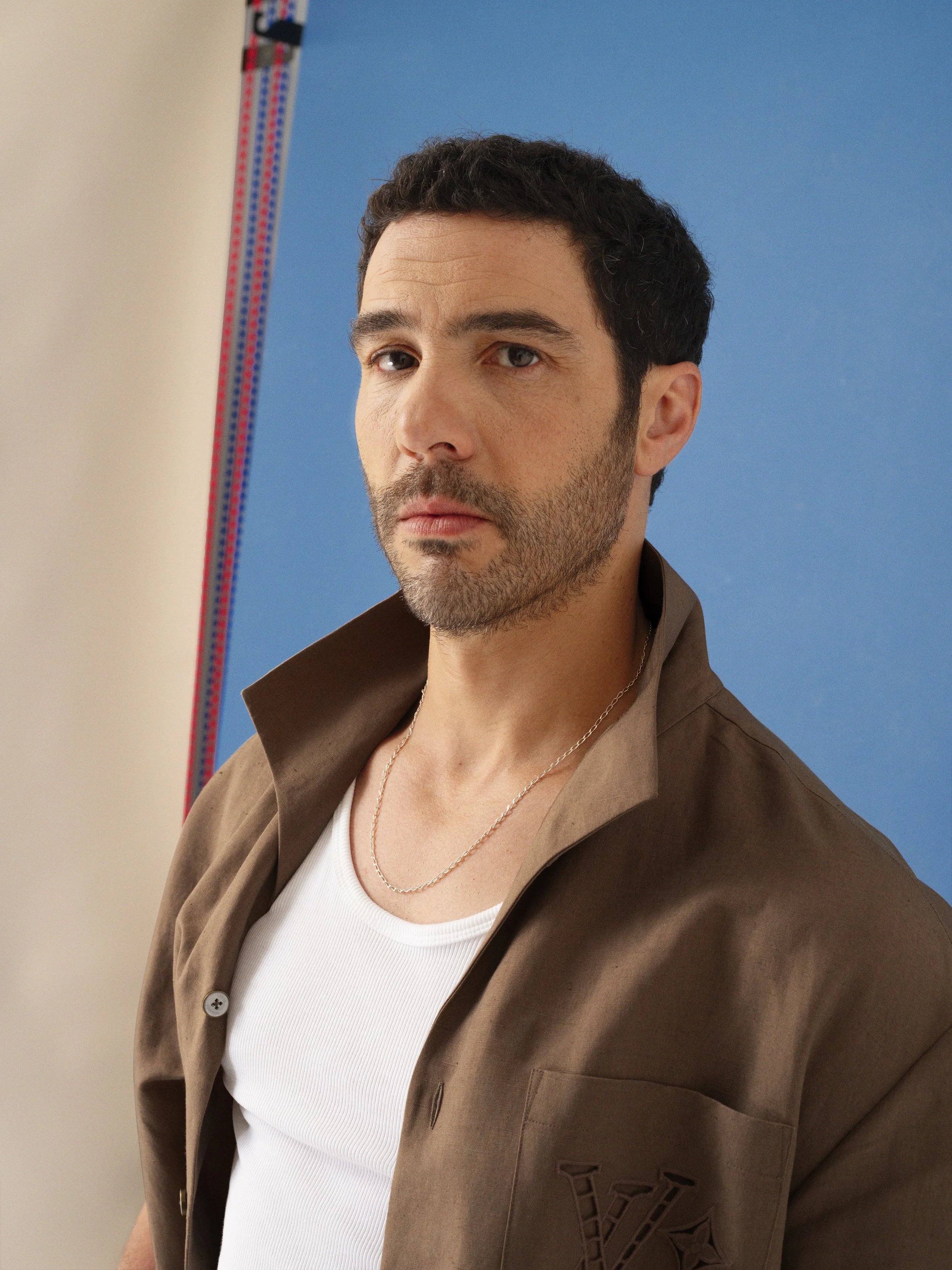 A man with dark hair and a beard looking serious, wearing a white tank top and a brown shirt with a collar, standing in front of a blue background.