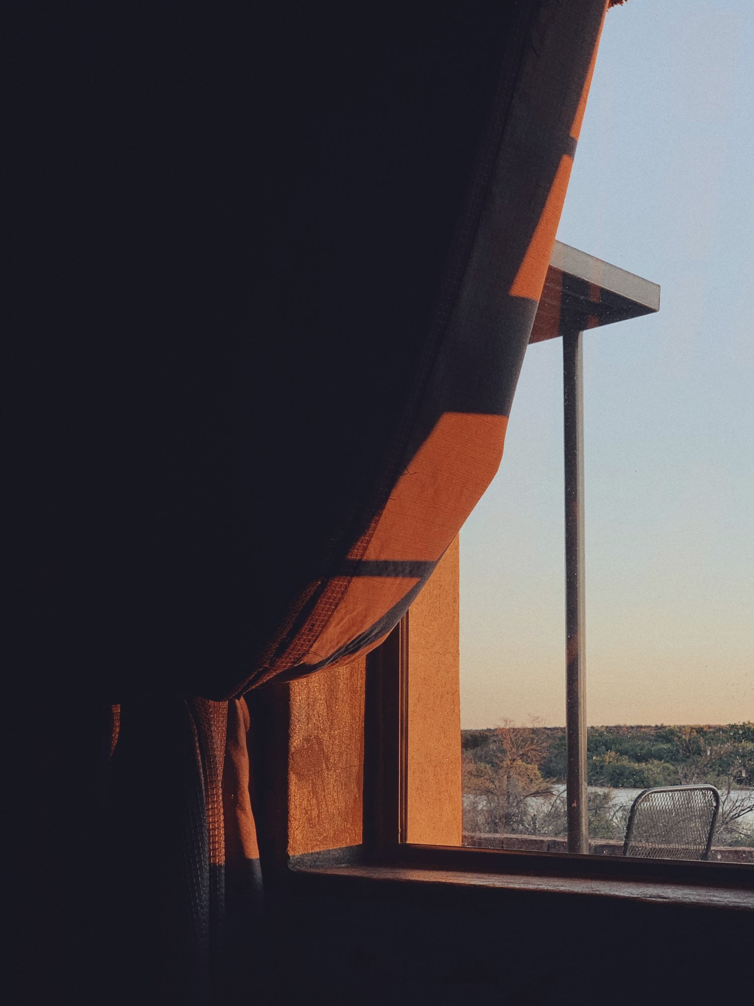 Sunlight coming through a window, illuminating a room with a curtain, view of trees and outdoor furniture outside.