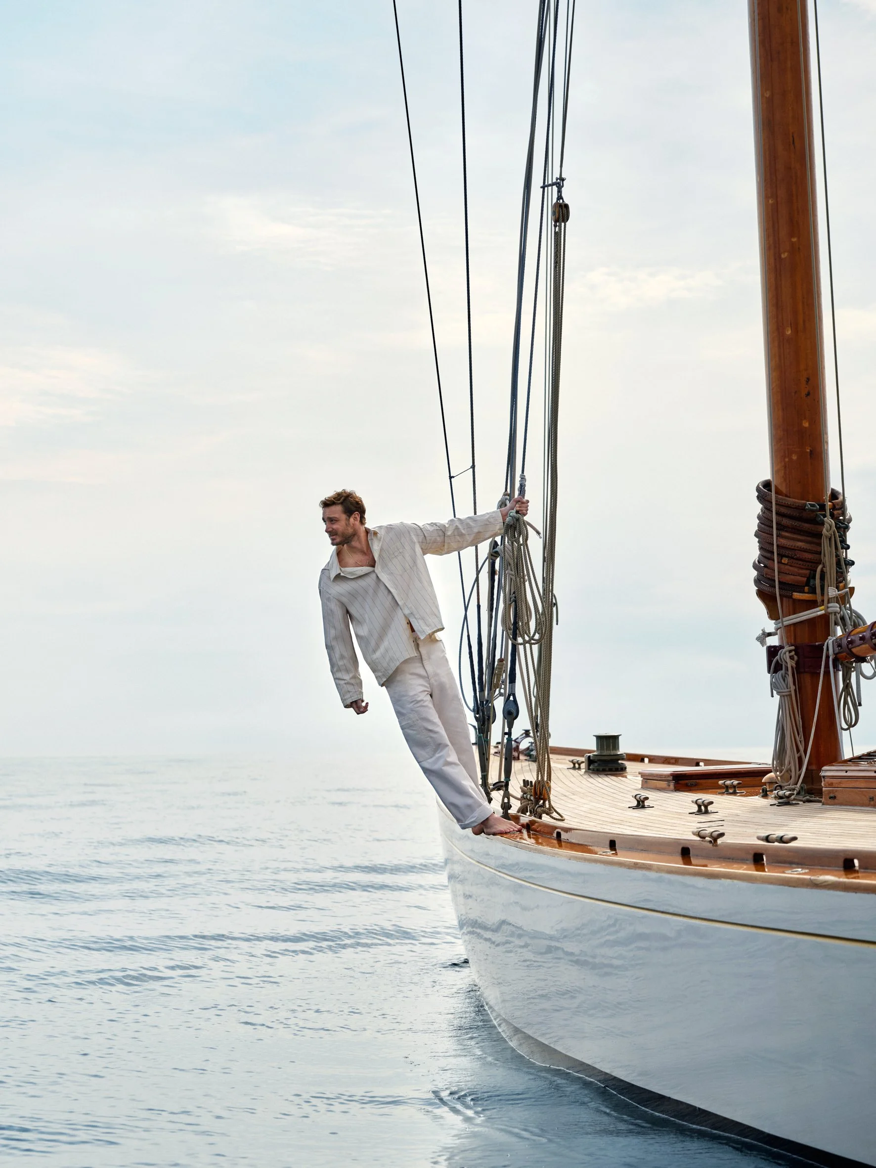 A man in pajamas leaning over the bow of a sailboat, holding onto the rigging, with calm ocean waters and a cloudy sky in the background.