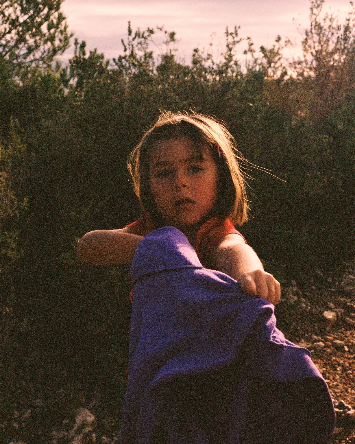 A young girl with shoulder-length brown hair, wearing a purple jacket, sits outdoors at sunset surrounded by bushes and trees.