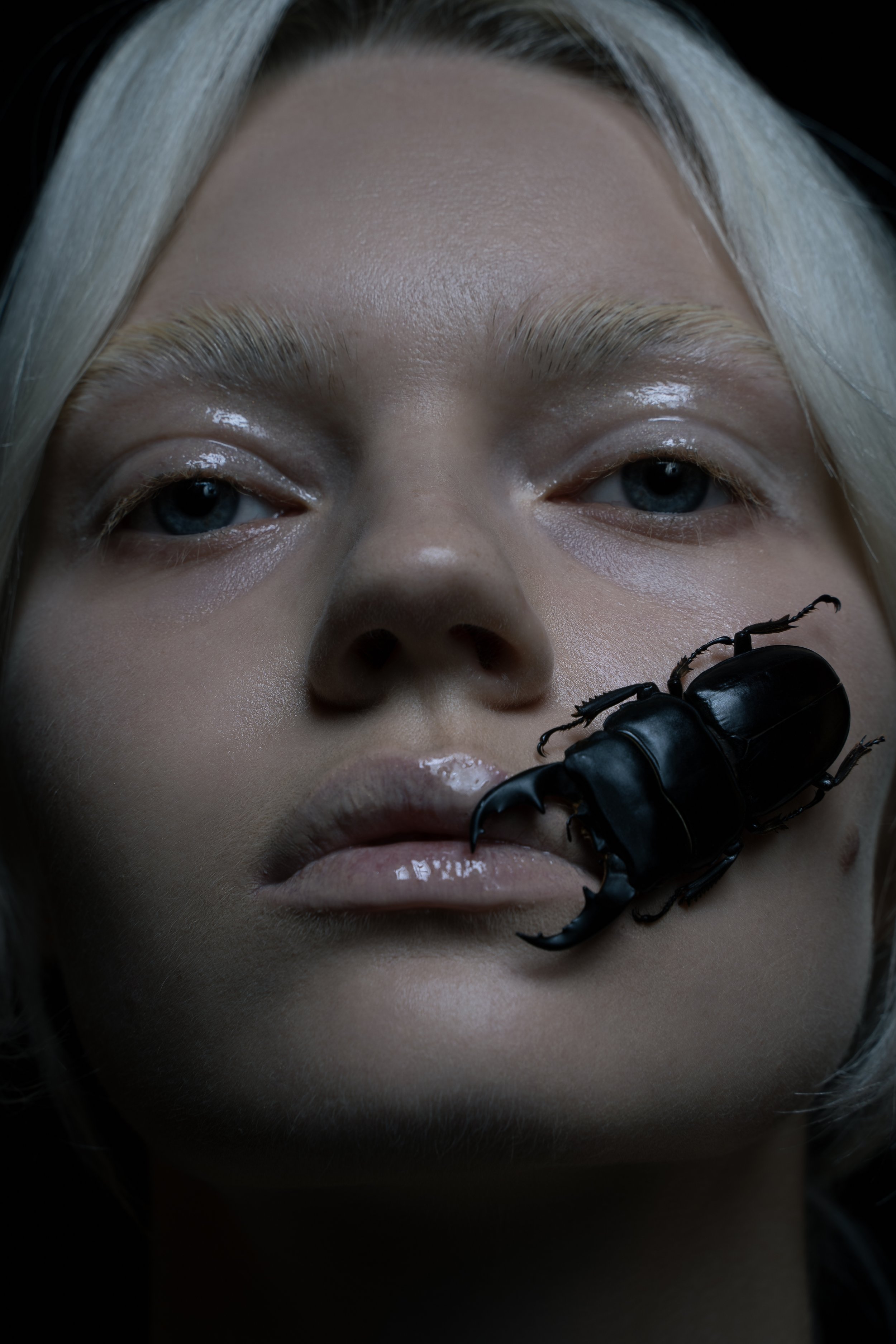 A close-up of a young woman with blonde hair and blue eyes, with a large black beetle on her lips.