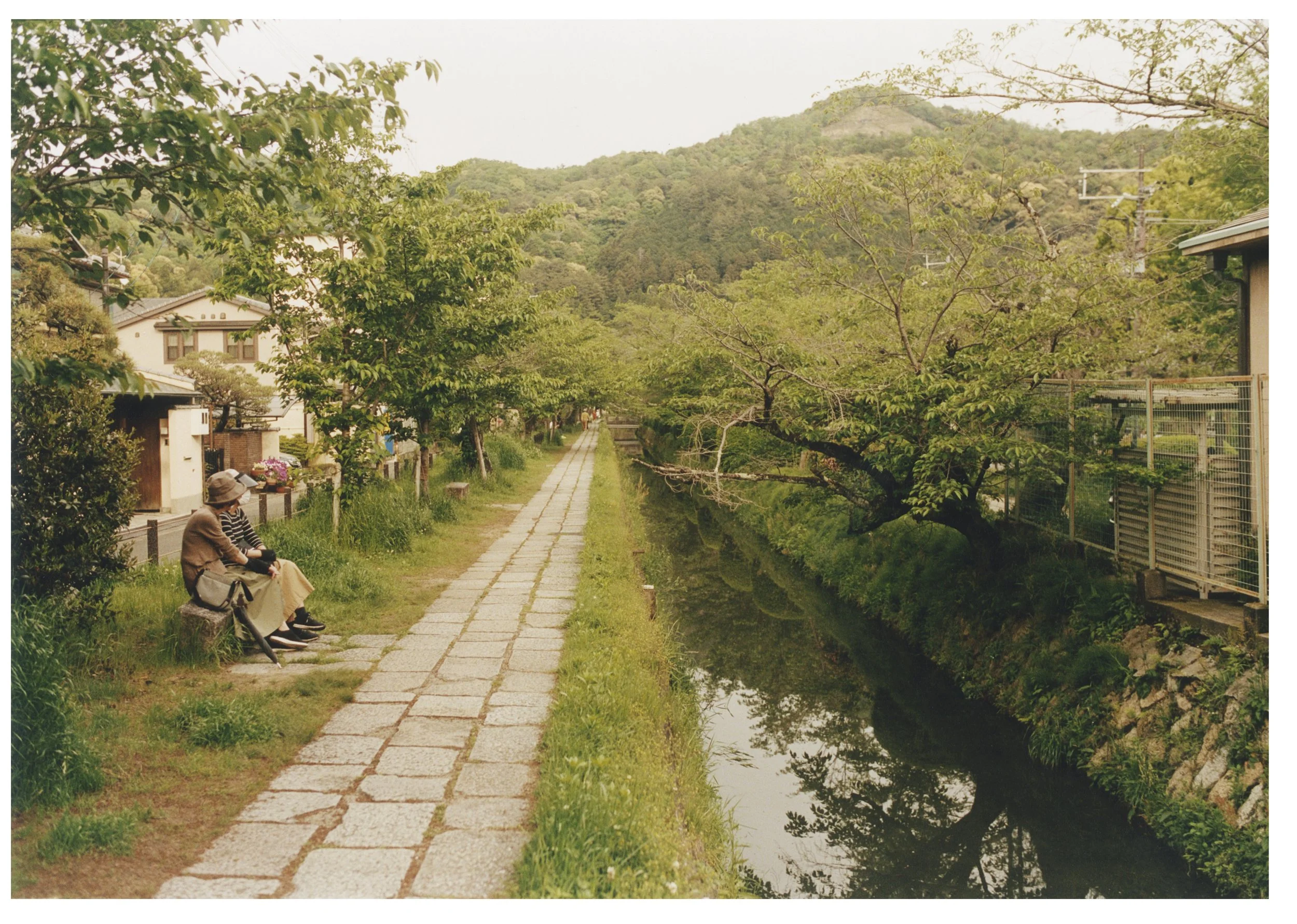 Two women sitting on a bench by a canal, surrounded by trees and houses, with distant hills in the background.